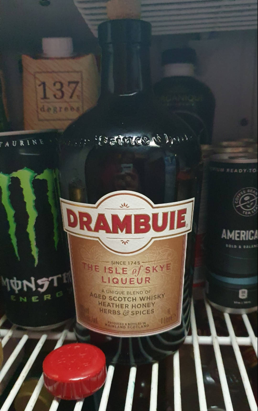 alcohol 桌布 壁紙 — 一瓶Drambuie利口酒放在冰箱架上，周圍是能量飲料和其他飲品。