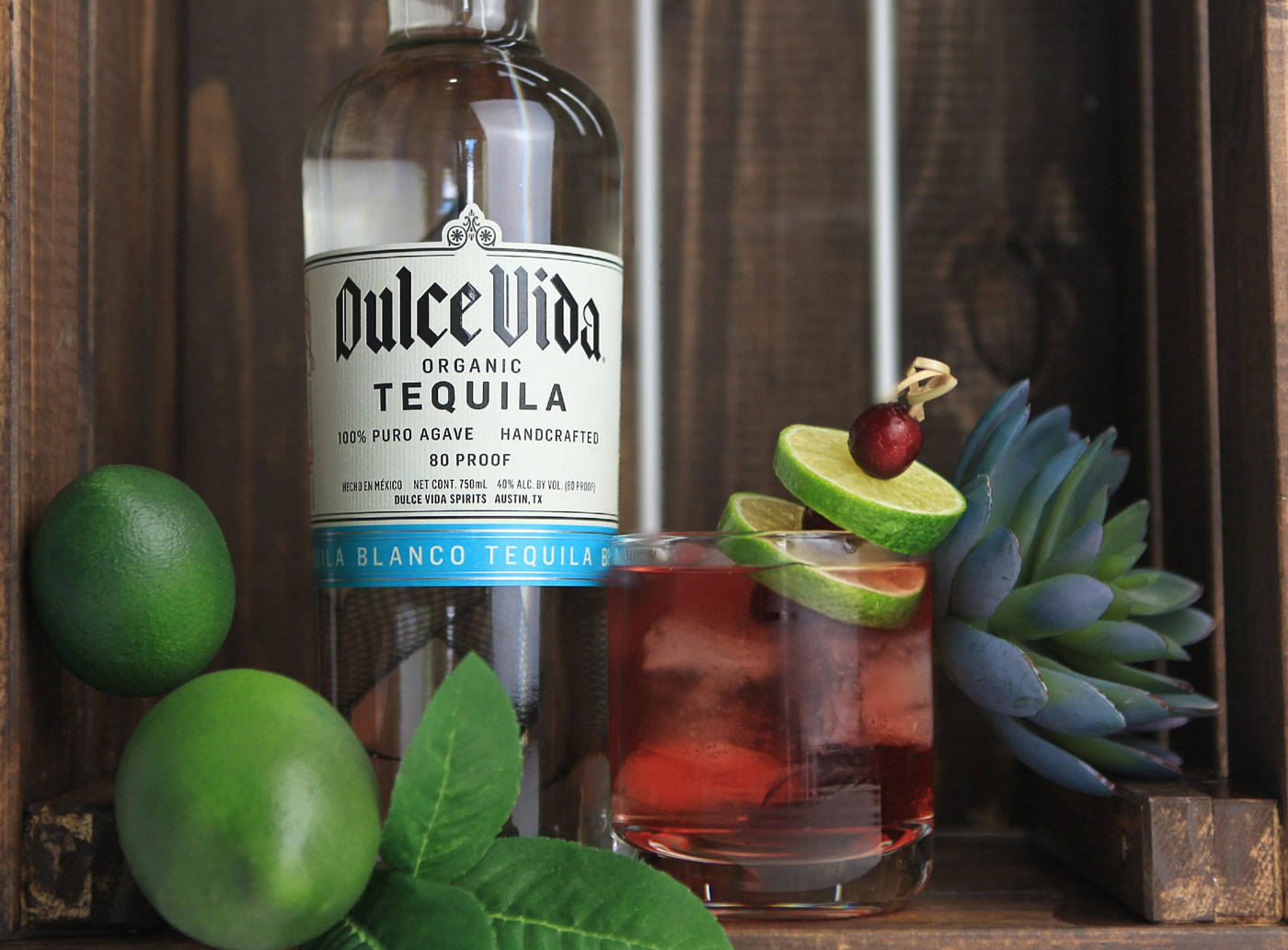 cocktail 桌布 壁紙 — 一瓶Dulce Vida龍舌蘭酒放在一個質樸的木箱中，旁邊是一杯用青檸和櫻桃裝飾的鮮紅色雞尾酒。