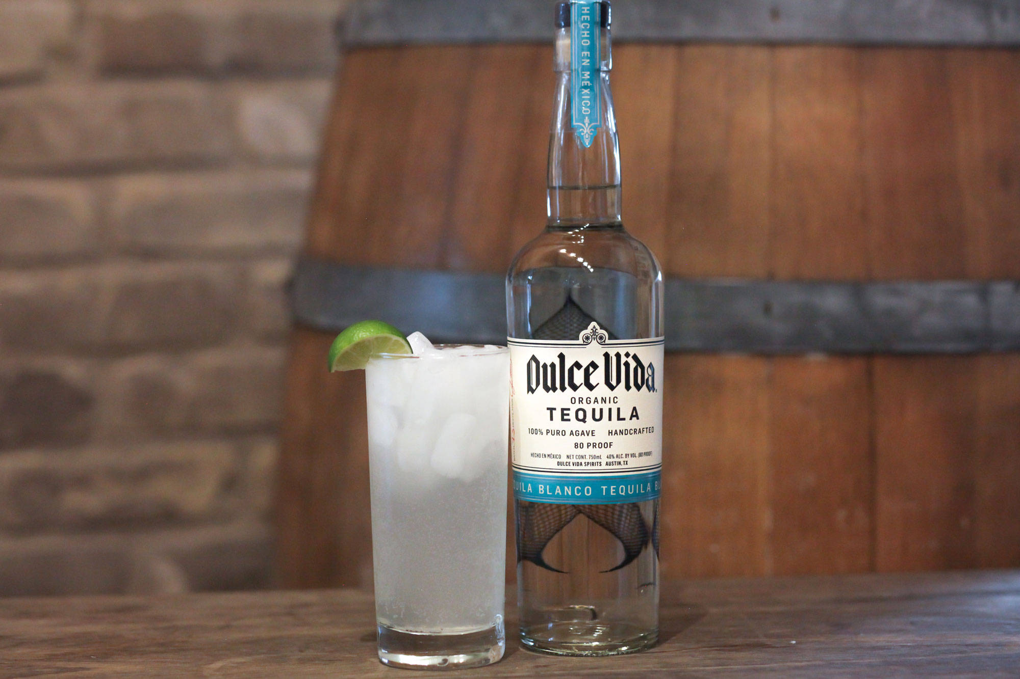 cocktail 桌布 壁紙 — 一瓶Dulce Vida龍舌蘭酒和一杯帶有萊姆角的冰凍雞尾酒，放在木桌上，背景是磚牆和木桶。