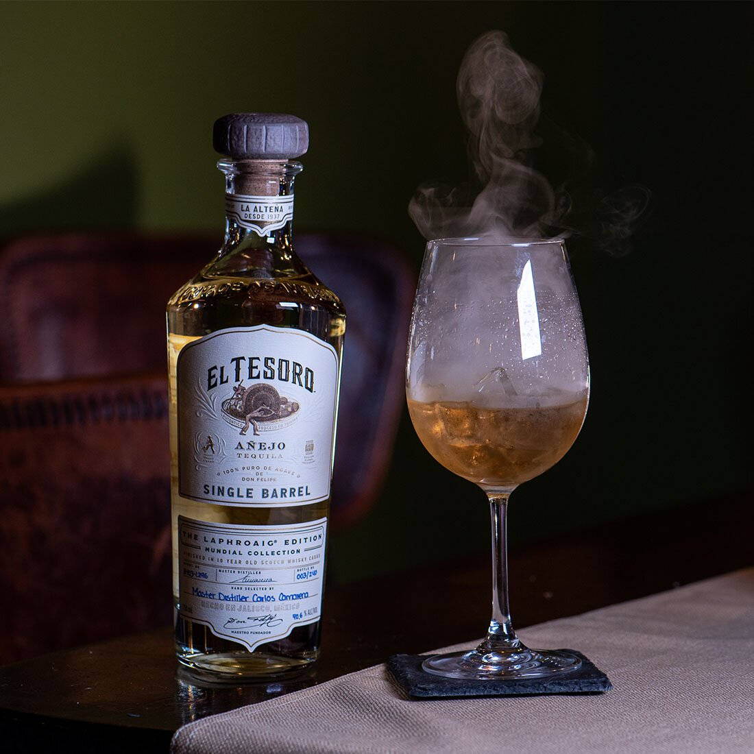 alcohol 桌布 壁紙 — 一瓶El Tesoro Añejo龍舌蘭酒放在一個有紋理的桌布上，旁邊有一杯冰凍龍舌蘭酒，煙霧裊裊升起。
