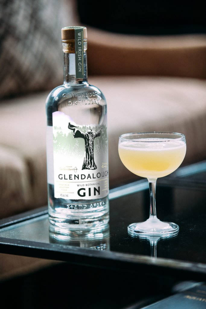 cocktail 桌布 壁紙 — 一瓶Glendalough Wild Irish Gin和一杯泡沫雞尾酒優雅地放在一張反光的黑色桌子上，旁邊是一張豪華沙發。