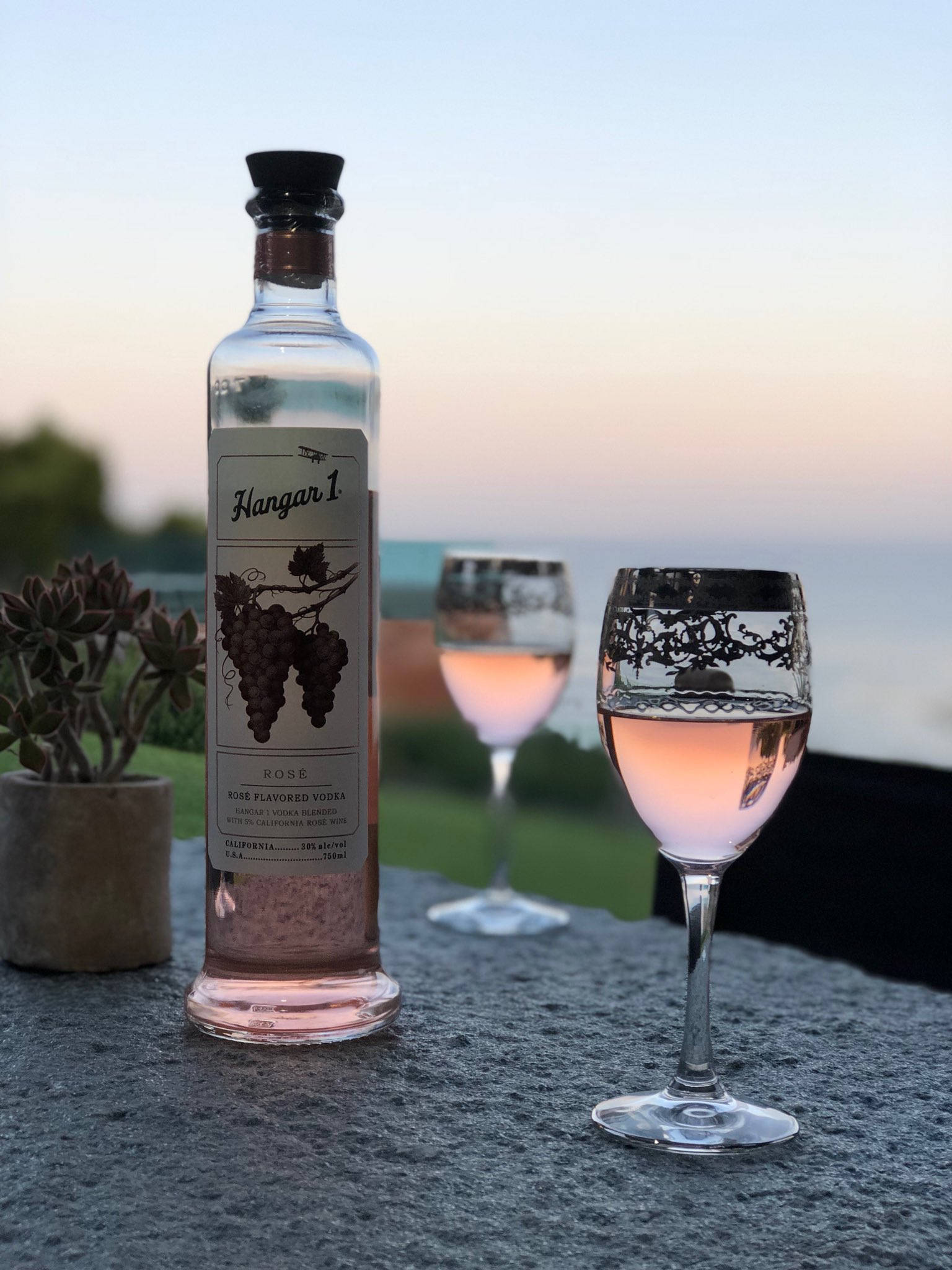 bottle 桌布 壁紙 — 一瓶Hangar 1玫瑰伏特加和兩個酒杯放在石檯上，俯瞰日落時分寧靜的海洋。