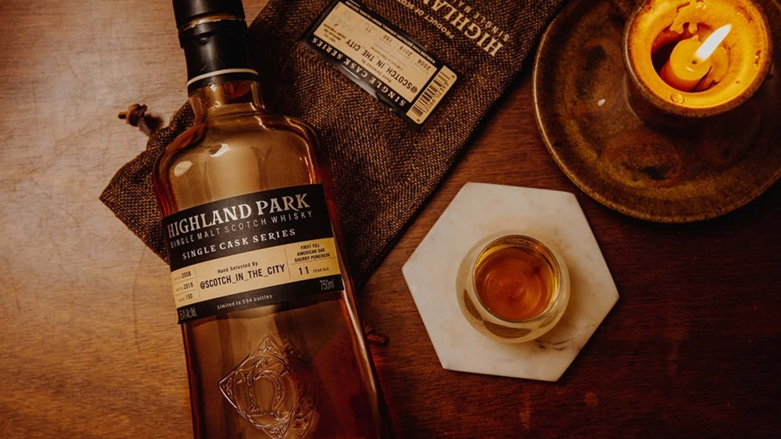 booze 桌布 壁紙 — 一瓶Highland Park威士忌放在一個質樸的木桌上，旁邊有一根點燃的蠟燭和一杯琥珀色的液體。