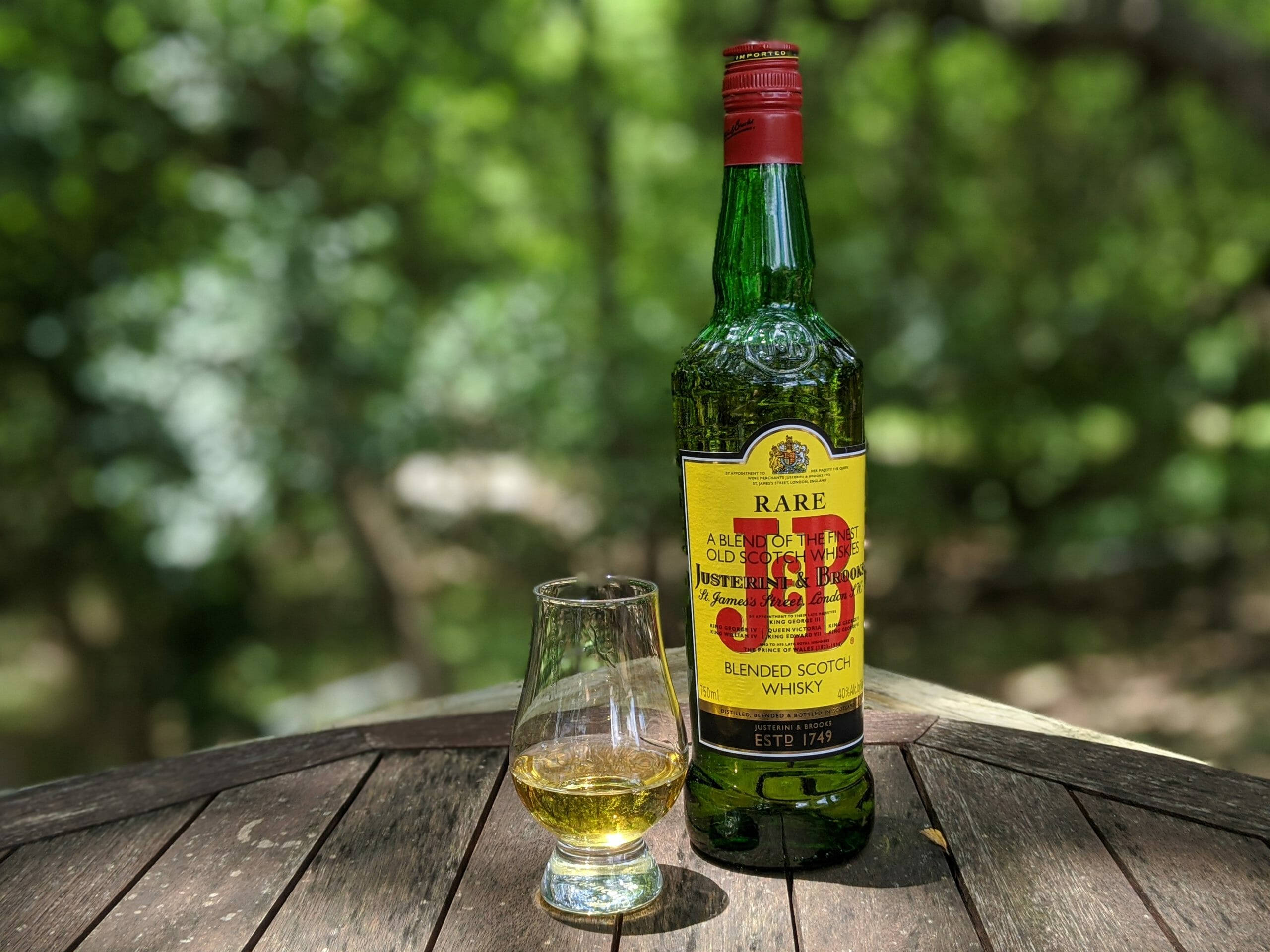 drinks 桌布 壁紙 — 一瓶J&B Rare威士忌和一個裝滿的品酒杯放在戶外一張風化的木桌上，背景是柔焦的茂密綠葉。
