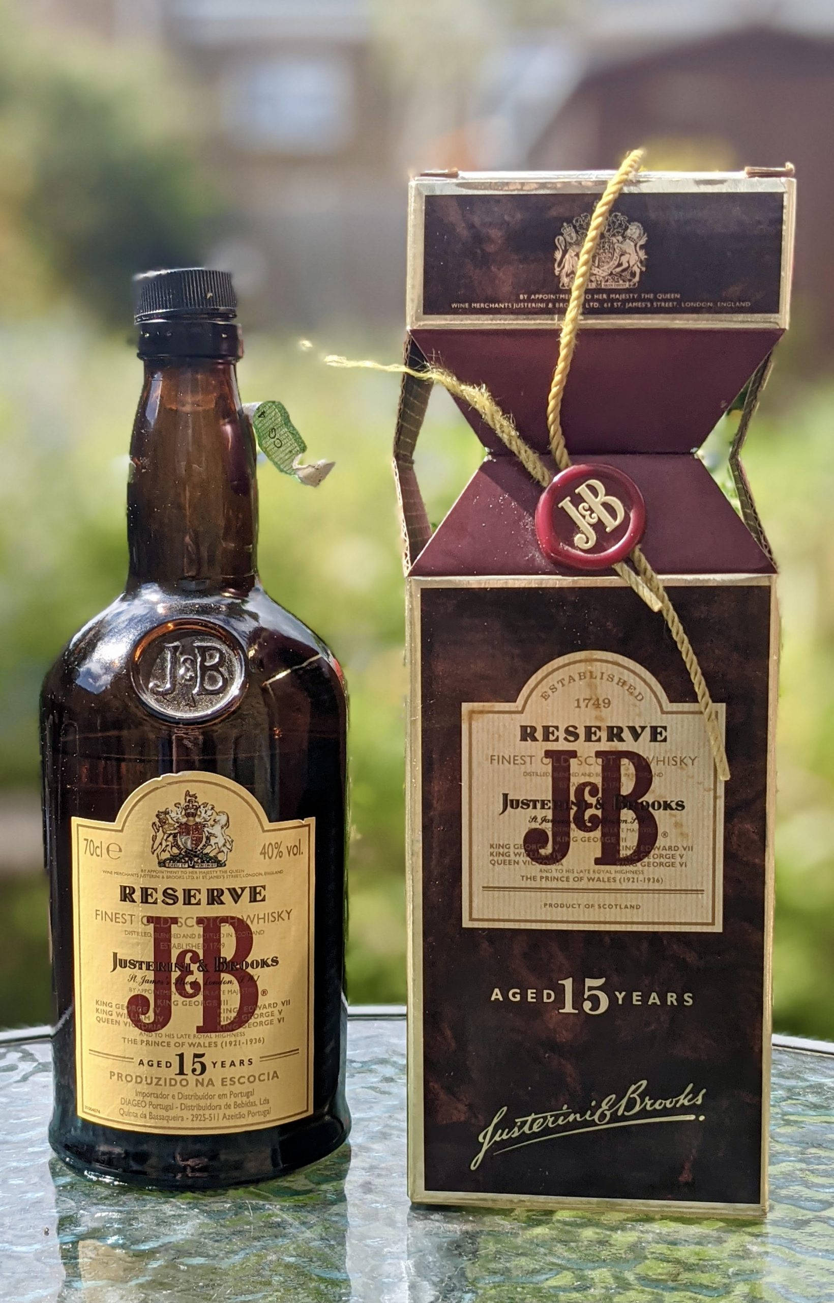 drinks 桌布 壁紙 — 一瓶J&B Reserve 15年威士忌及其優雅的盒子放在玻璃桌上，背景是模糊的綠色植物。