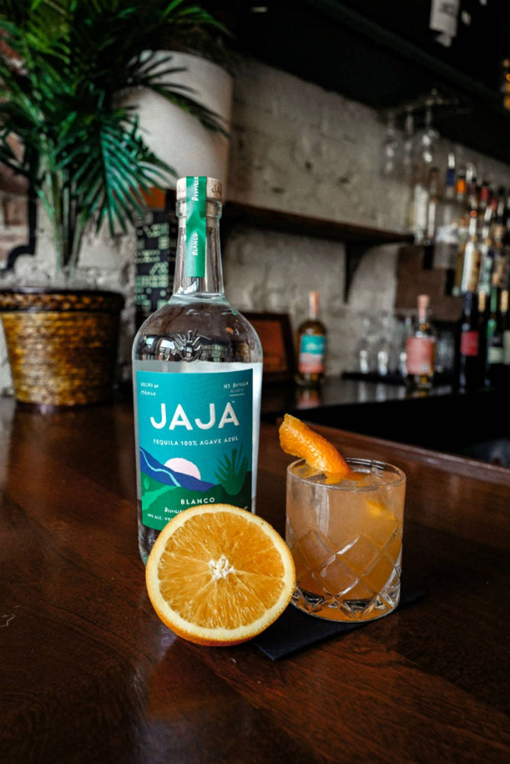 cocktail 桌布 壁紙 — 一瓶Jaja Blanco龍舌蘭酒放在深色木吧檯上，旁邊有柑橘雞尾酒和半個柳橙，背景有盆栽和放滿酒的架子。