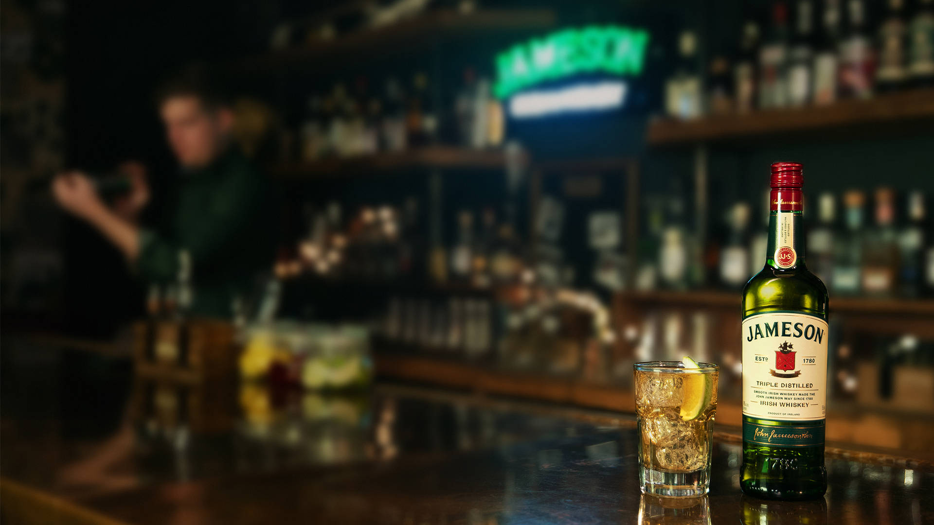 Bar 桌布 壁紙 — 一瓶Jameson愛爾蘭威士忌和一杯加冰和檸檬的玻璃杯放在拋光的吧檯上，背景中調酒師模糊，環境燈光溫暖。