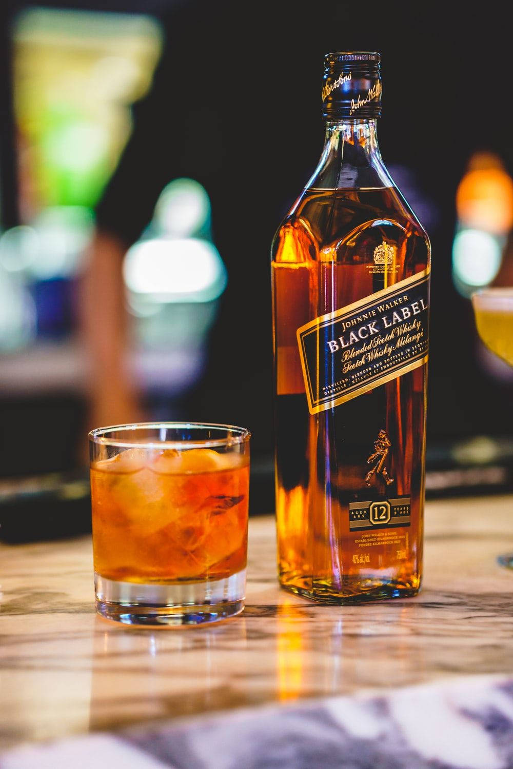 alcohol 桌布 壁紙 — 一瓶Johnnie Walker Black Label威士忌和一杯加冰的威士忌放在大理石吧檯上，背景是模糊的霓虹燈光。