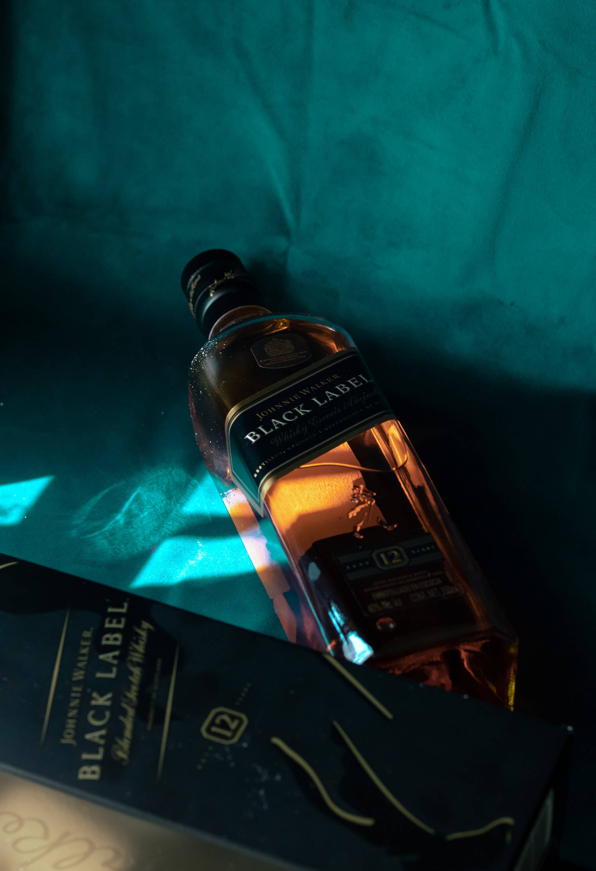 bottle 桌布 壁紙 — 一瓶Johnnie Walker Black Label威士忌酒瓶斜放在其盒子上，背景為青綠色布料，在戲劇性的燈光下。