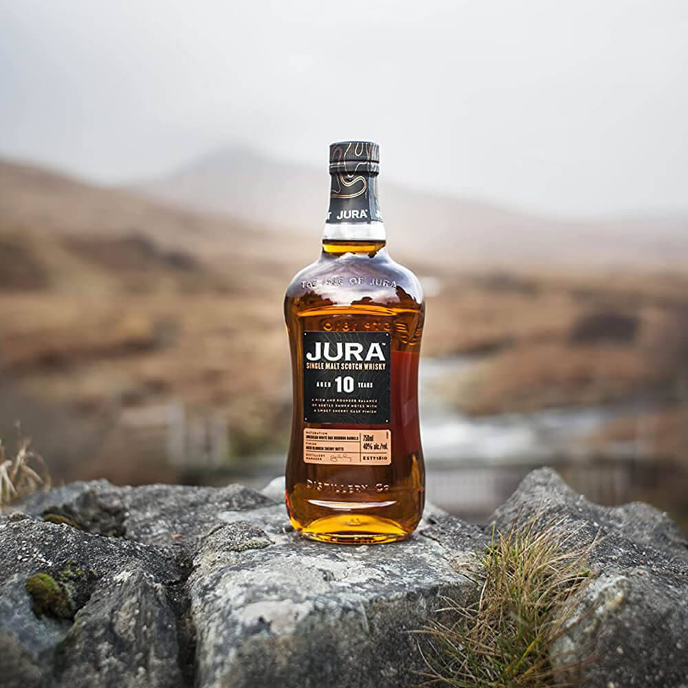 高清 桌布 壁紙 — 一瓶Jura 10年單一麥芽威士忌放在粗糙的灰色岩石上，背景是霧氣瀰漫的山丘。