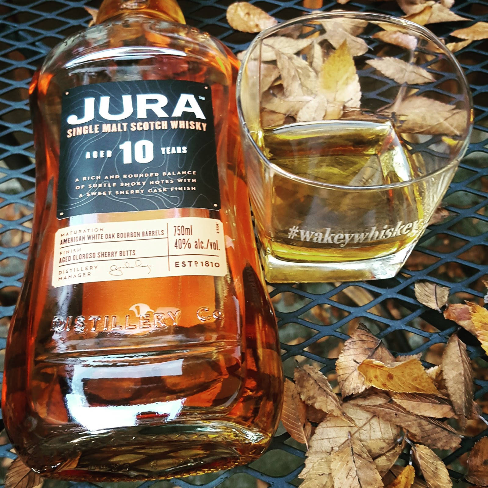 動態圖 桌布 壁紙 — 一瓶Jura 10年單一麥芽蘇格蘭威士忌放在一個金屬網桌上，旁邊有一個裝滿琥珀色液體的玻璃杯，周圍環繞著乾燥的秋葉。
