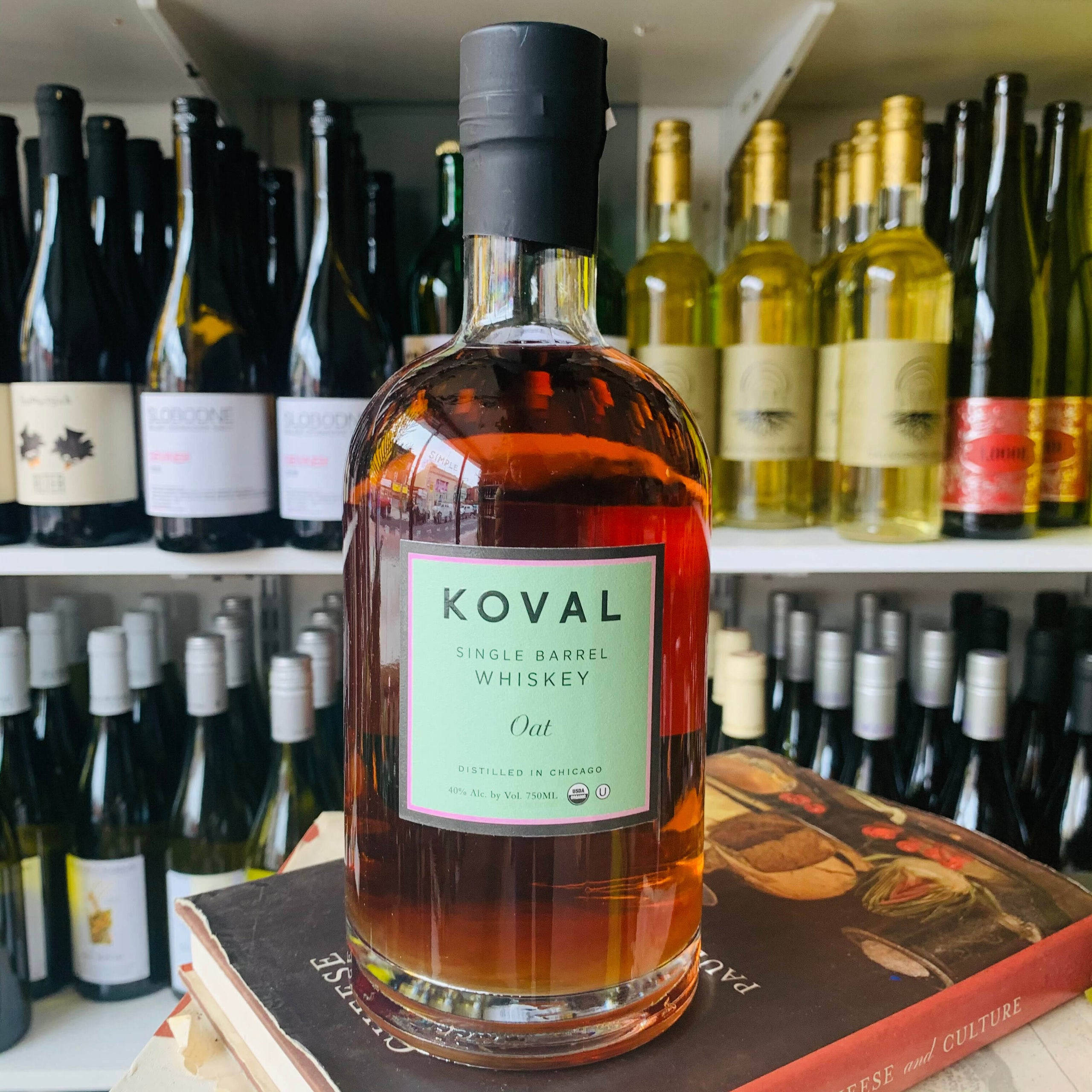 drinks 桌布 壁紙 — 一瓶Koval單桶燕麥威士忌醒目地放在一本書上，書本置於擺滿酒瓶的架子上，店內光線充足。