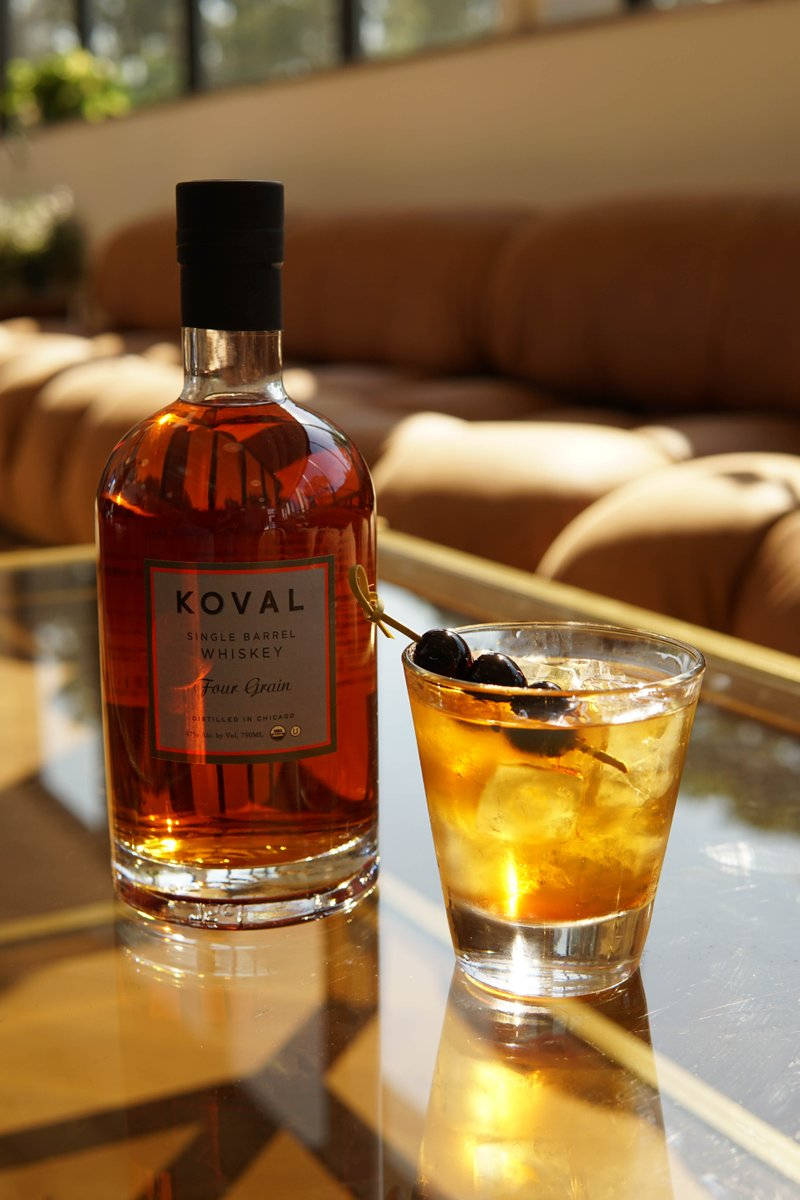 cocktail 桌布 壁紙 — 一瓶Koval威士忌和一杯帶橄欖的雞尾酒放在玻璃桌上，沐浴在溫暖的陽光中，旁邊是一張棕褐色沙發。