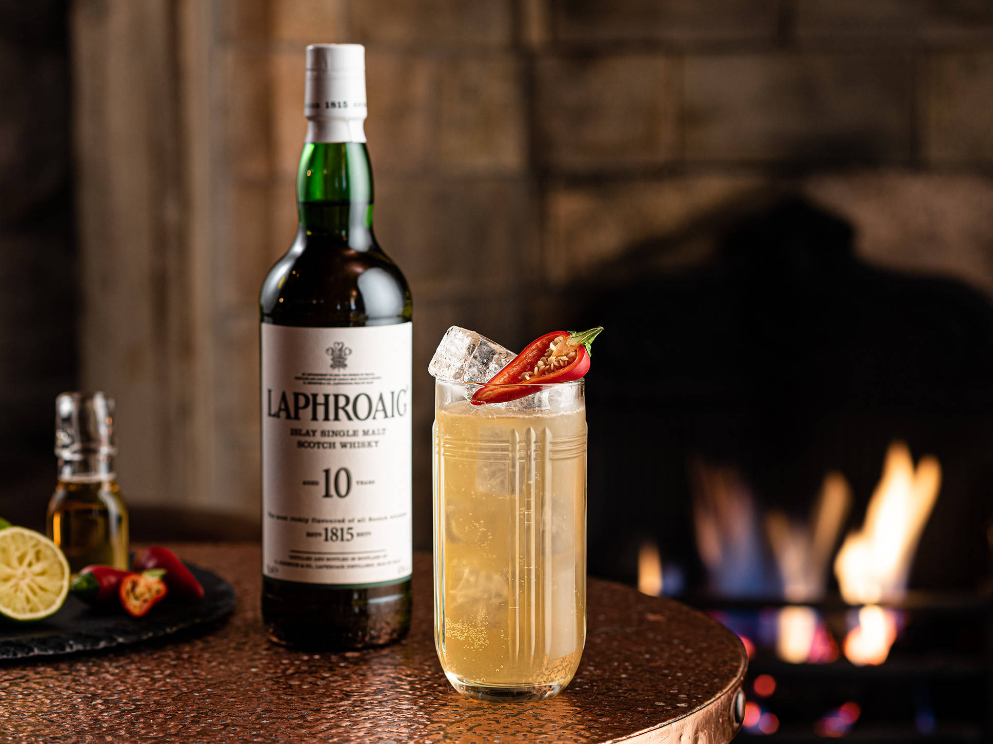 Best 桌布 壁紙 — 一瓶Laphroaig 10年威士忌和帶有辣椒裝飾的辛辣雞尾酒，放在銅桌上，前面是劈啪作響的壁爐。
