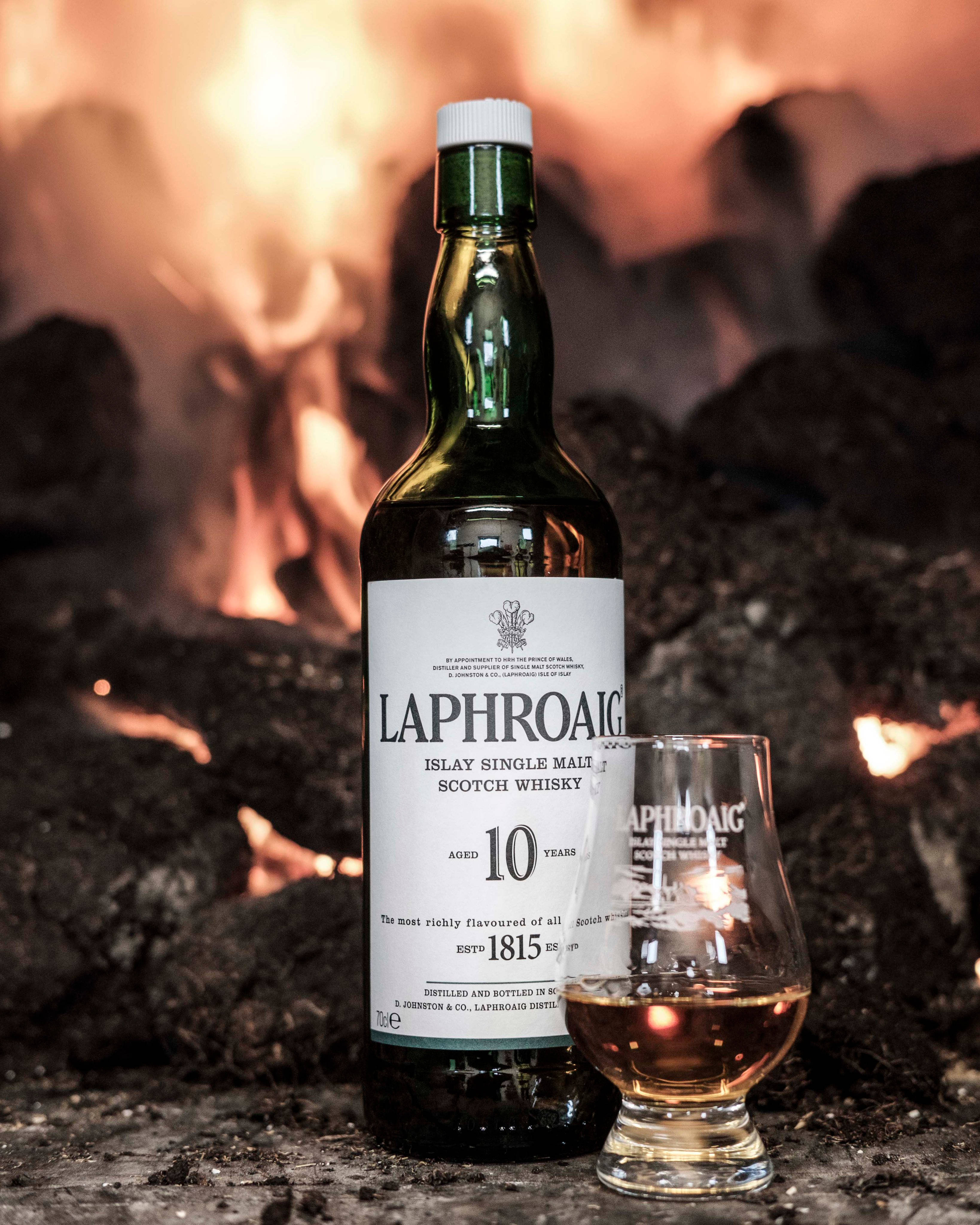 iPhone 桌布 壁紙 — 一瓶Laphroaig 10年艾雷島單一麥芽蘇格蘭威士忌和一個裝滿酒的杯子放在熊熊烈火前，火焰中帶有發光的餘燼。