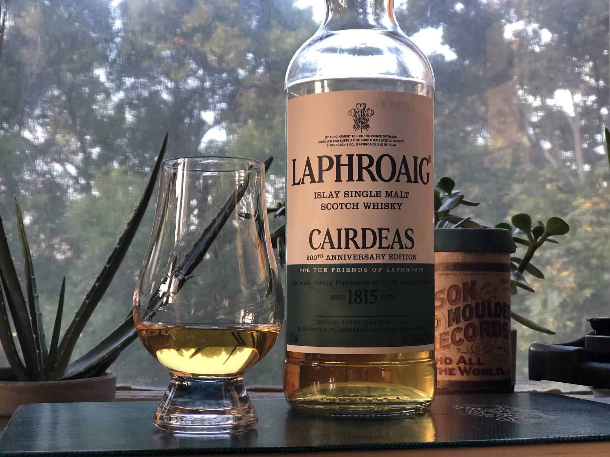 安卓 桌布 壁紙 — 一瓶Laphroaig Cairdeas威士忌和一個裝滿金色液體的品酒杯放在靠近窗戶的深色墊子上，窗外有綠色植物。