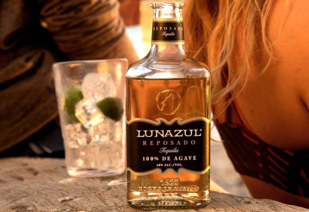 alcohol 桌布 壁紙 — 一瓶Lunazul Reposado龍舌蘭酒放在石桌上，旁邊有一杯加冰和萊姆的酒杯，一位女士靠在附近。