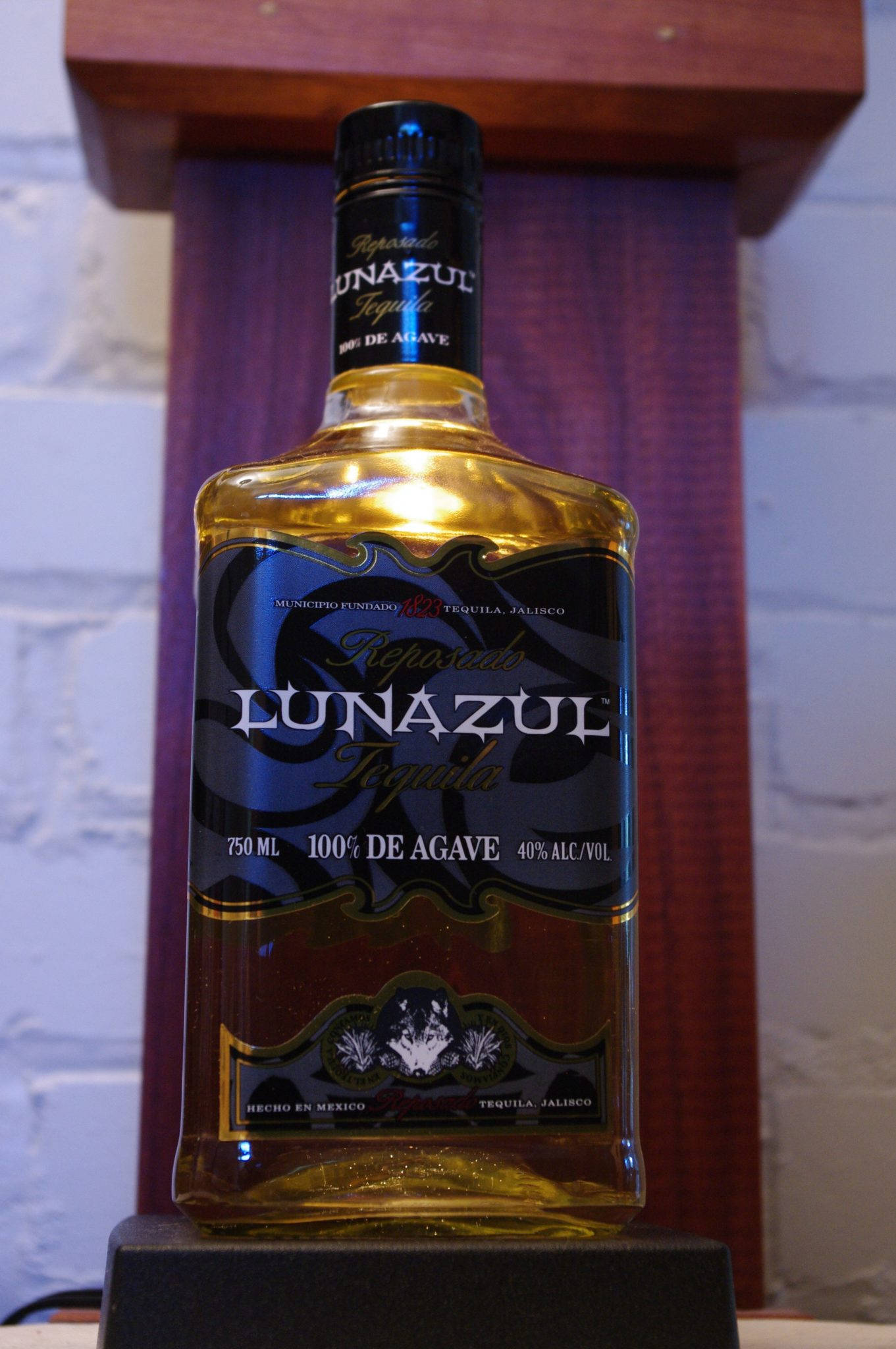 alcohol 桌布 壁紙 — 一瓶Lunazul Reposado龍舌蘭酒在白色磚牆和木架前的深色支架上溫暖地發光。