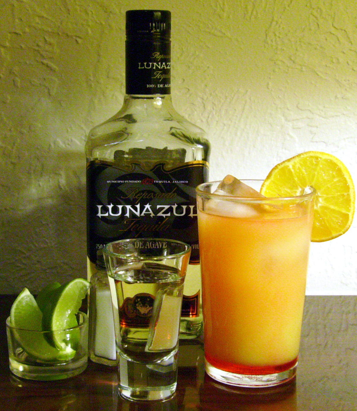 alcohol 桌布 壁紙 — 一瓶Lunazul龍舌蘭酒放在一個高身橙色雞尾酒杯旁邊，杯中有冰塊和萊姆裝飾，背景是紋理牆壁。