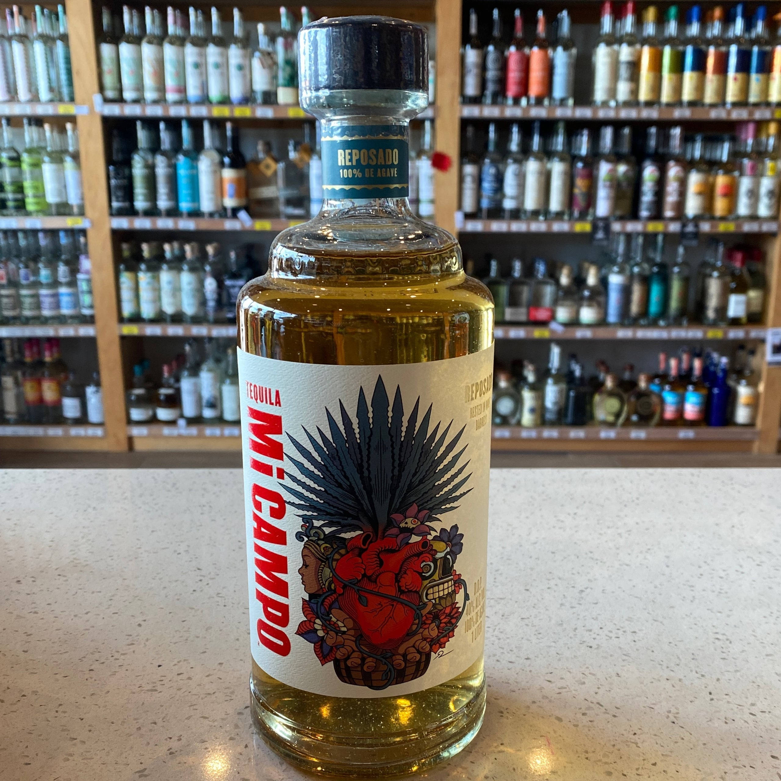 bottle 桌布 壁紙 — 一瓶Mi Campo reposado龍舌蘭酒，帶有鮮豔的骷髏頭和龍舌蘭標籤，放在一個有斑點的檯面上，前面是擺滿酒瓶的架子。