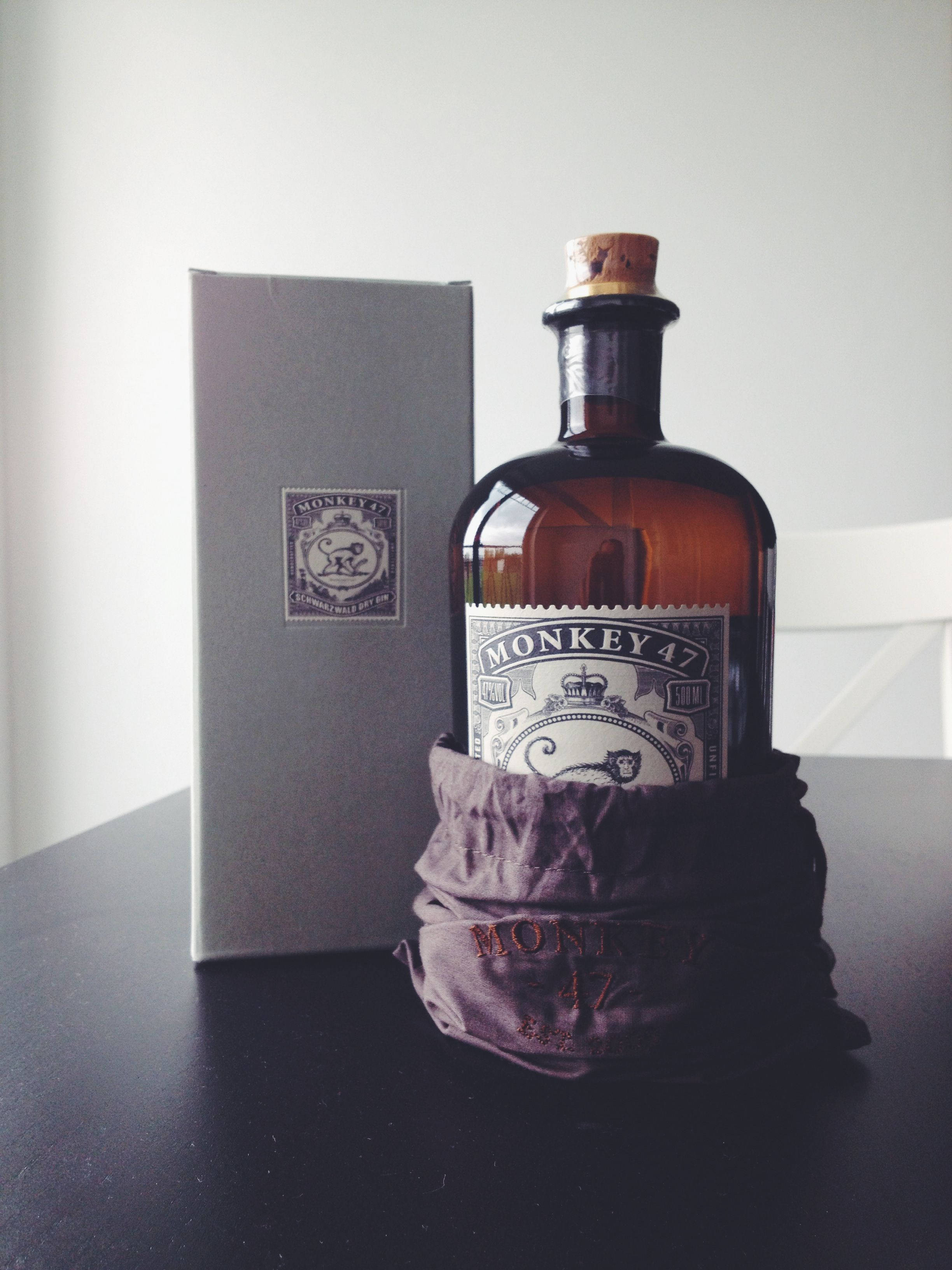 飲料 桌布 壁紙 — 一瓶Monkey 47琴酒優雅地放在天鵝絨套中，旁邊是其灰色盒子，置於深色桌上。