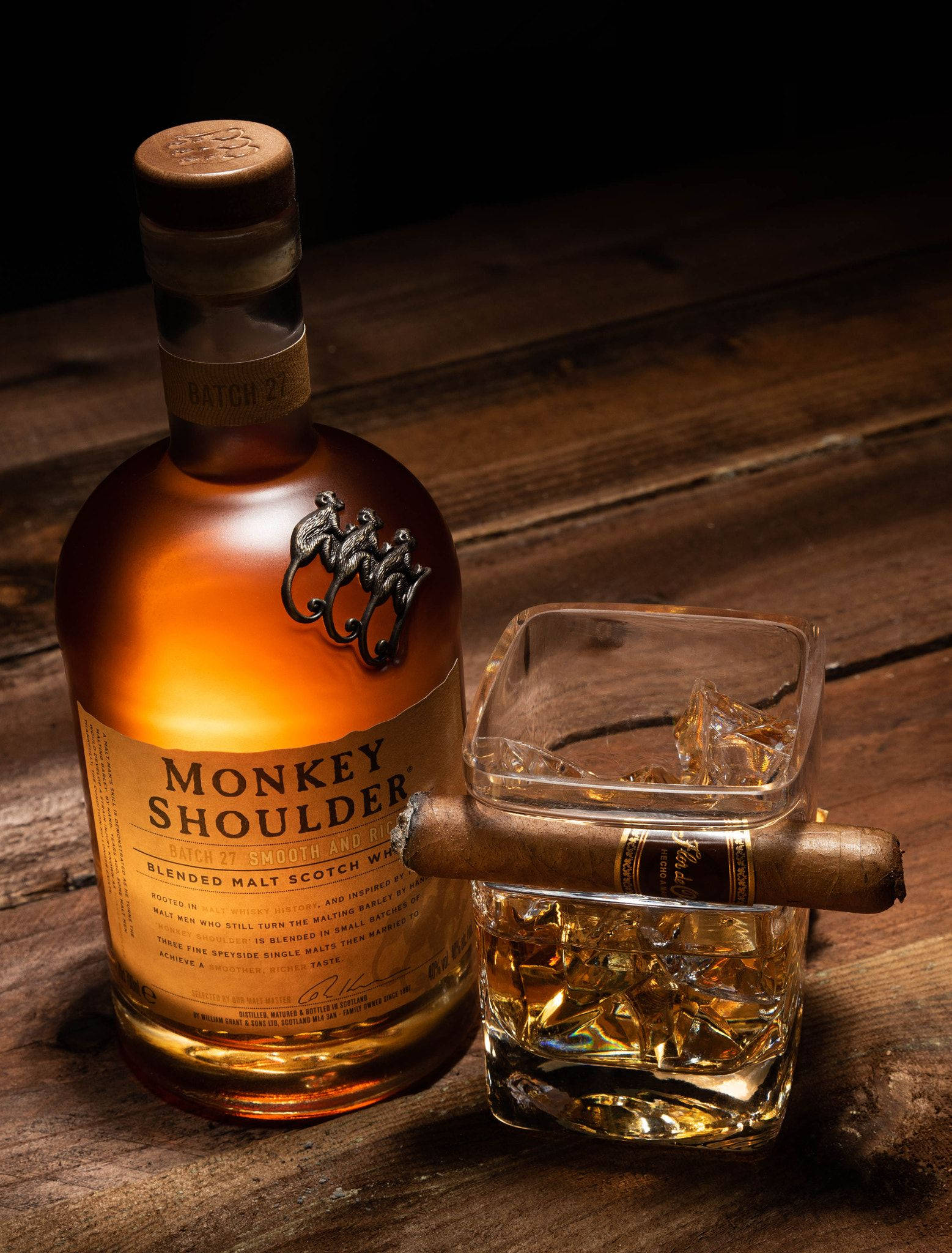 飲料 桌布 壁紙 — 一瓶Monkey Shoulder威士忌、一杯加冰的威士忌和一支雪茄放在深色木桌上，溫暖的燈光下。