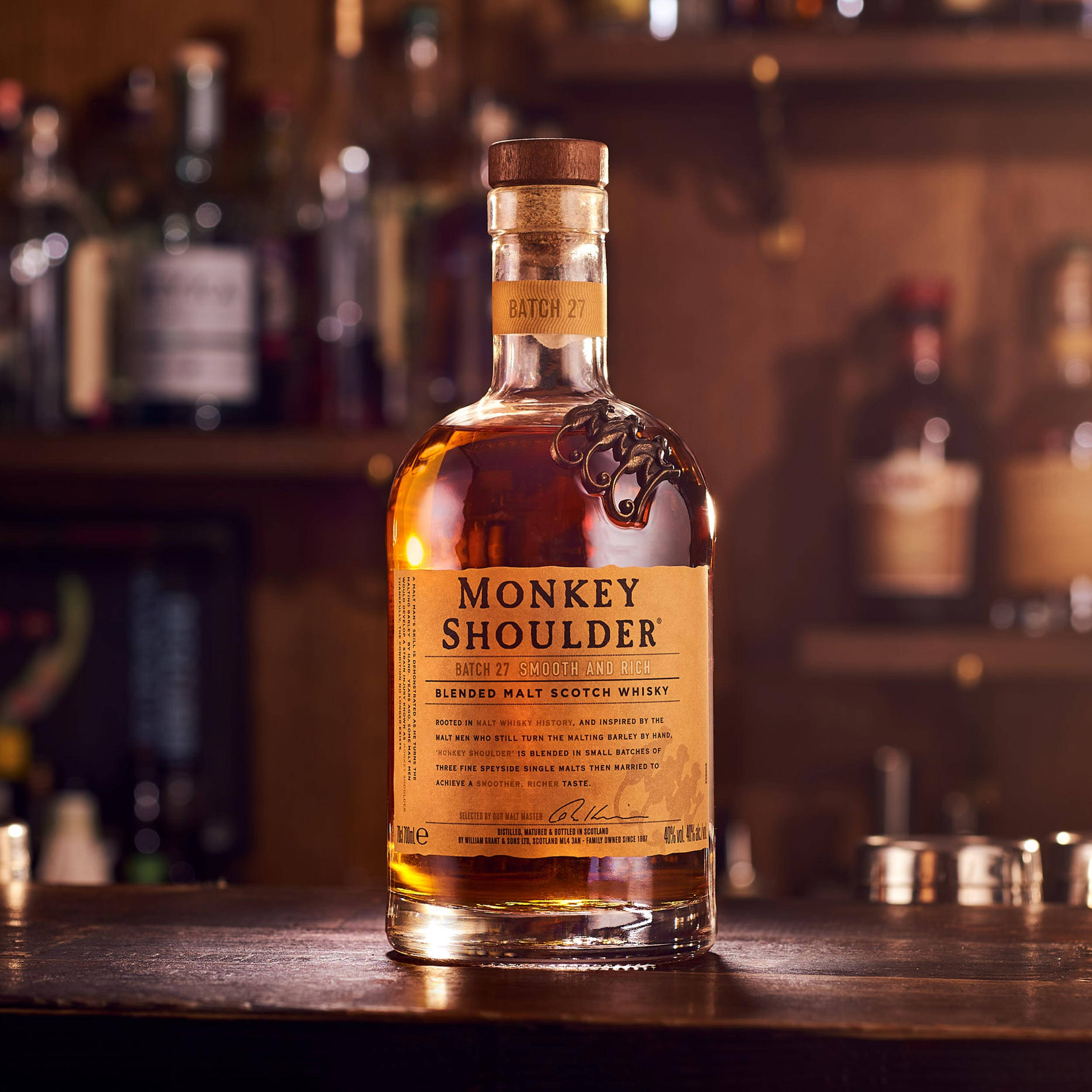 alcohol 桌布 壁紙 — 一瓶Monkey Shoulder威士忌在木製吧台上溫暖地發光，背景是模糊的酒架。