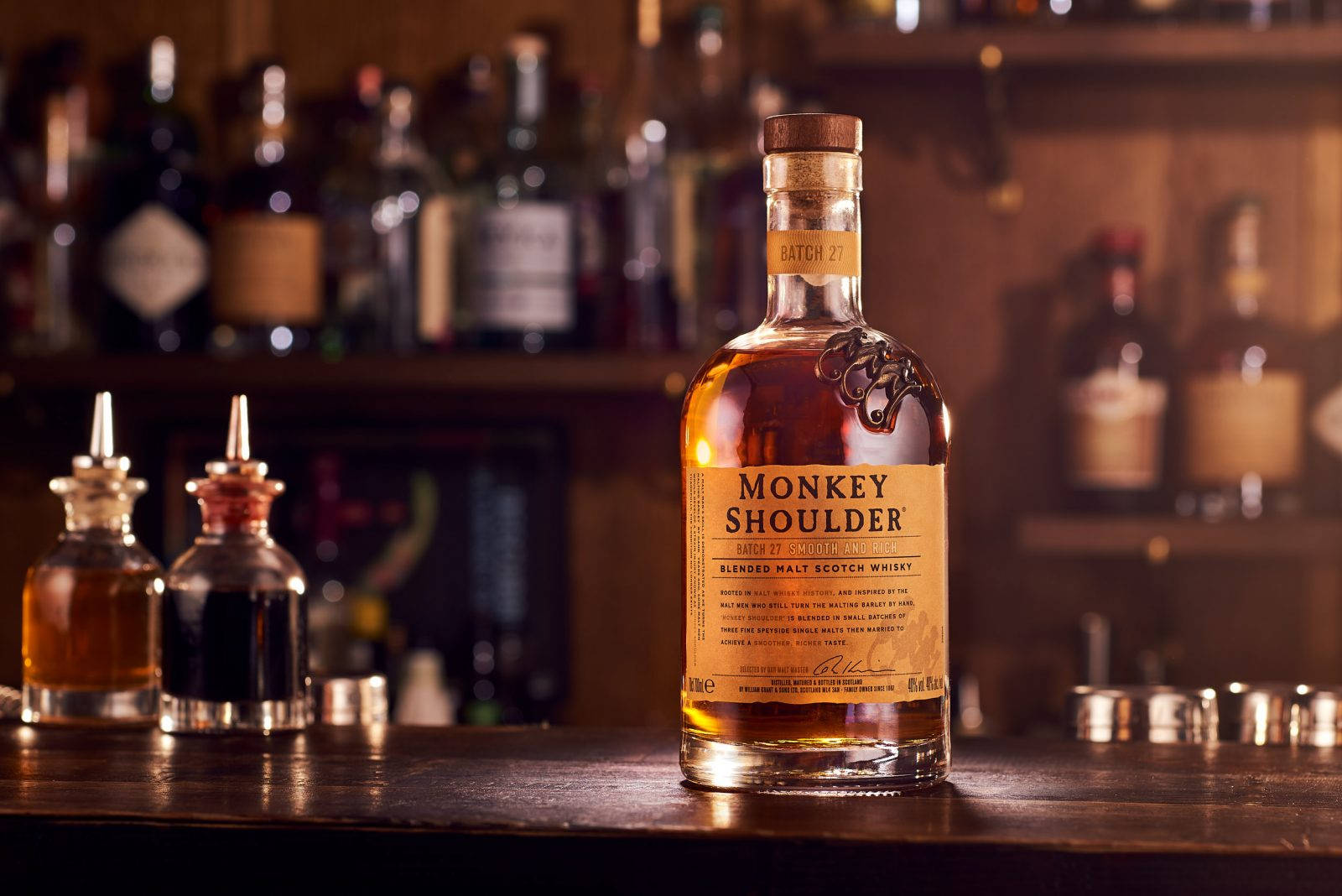 alcohol 桌布 壁紙 — 一瓶Monkey Shoulder威士忌在昏暗的酒吧中，於木製吧檯上溫暖地發光，周圍環繞著調酒器和其他烈酒。