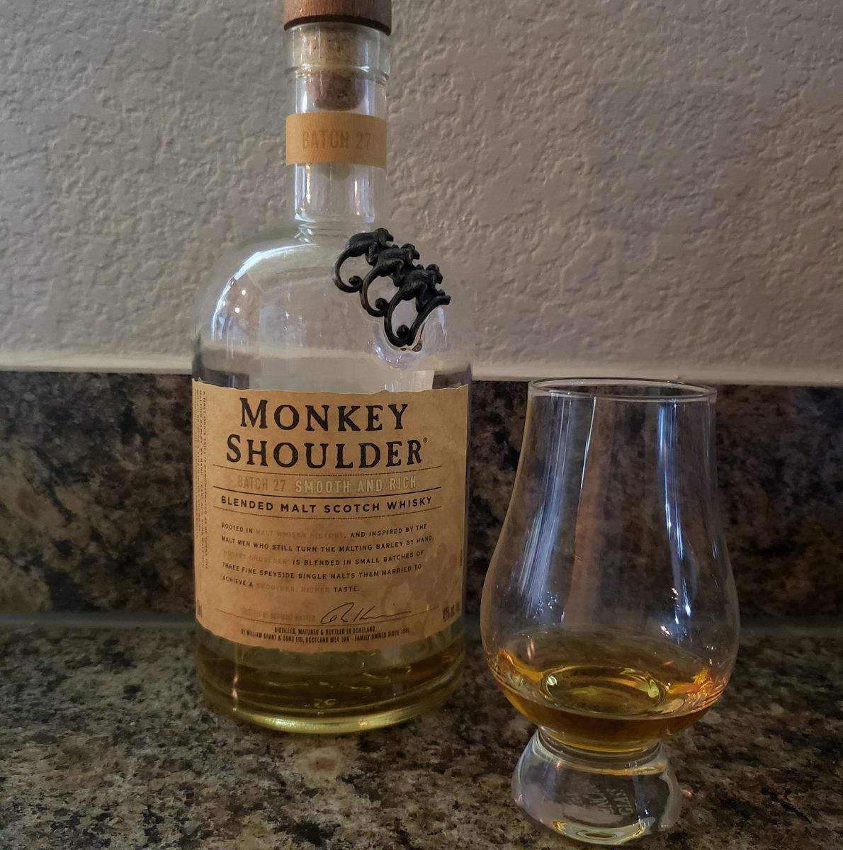 alcohol 桌布 壁紙 — 一瓶Monkey Shoulder威士忌和一個品酒杯放在花崗岩檯面上，背景是紋理牆壁。