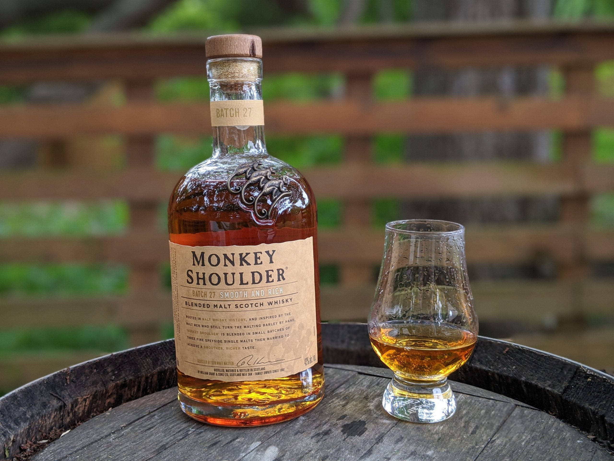 alcohol 桌布 壁紙 — 一瓶Monkey Shoulder威士忌和一杯琥珀色液體放在一個風化的木桶頂部，背景是模糊的木柵欄和綠色植物。