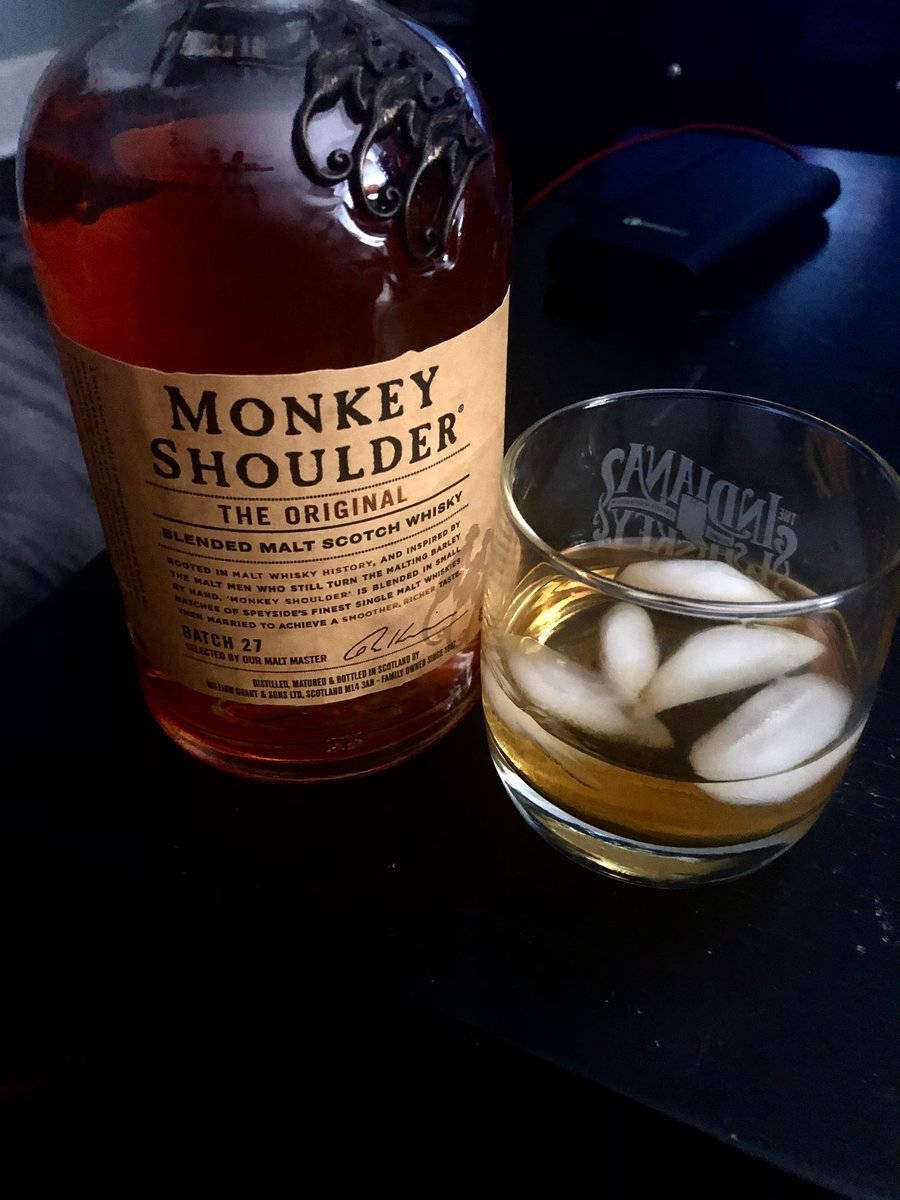 飲料 桌布 壁紙 — 一瓶Monkey Shoulder威士忌和一杯加冰的威士忌放在昏暗燈光下的深色表面上。