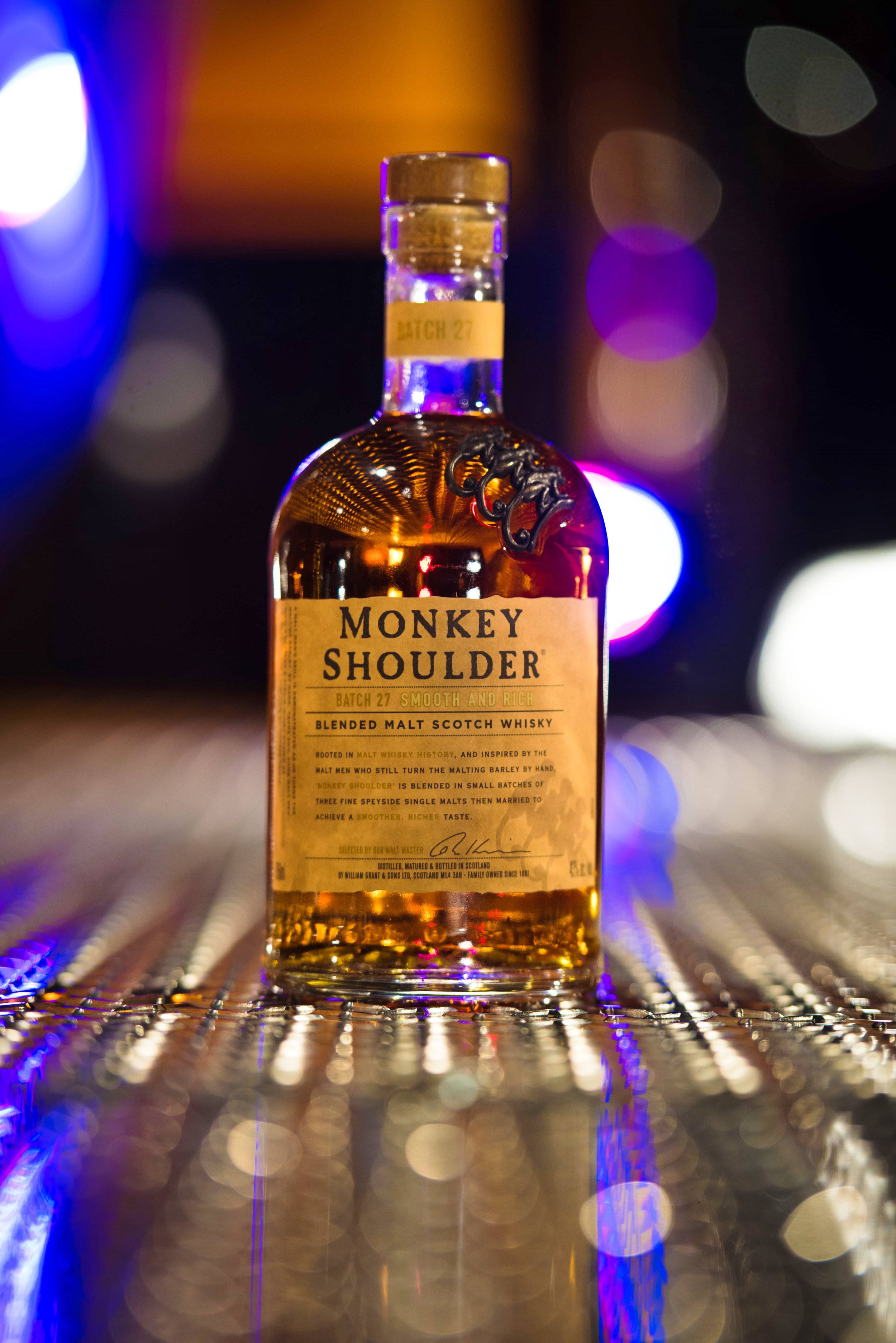 alcohol 桌布 壁紙 — 一瓶Monkey Shoulder威士忌在反射的吧檯表面上，於色彩繽紛的散景燈光下閃耀。