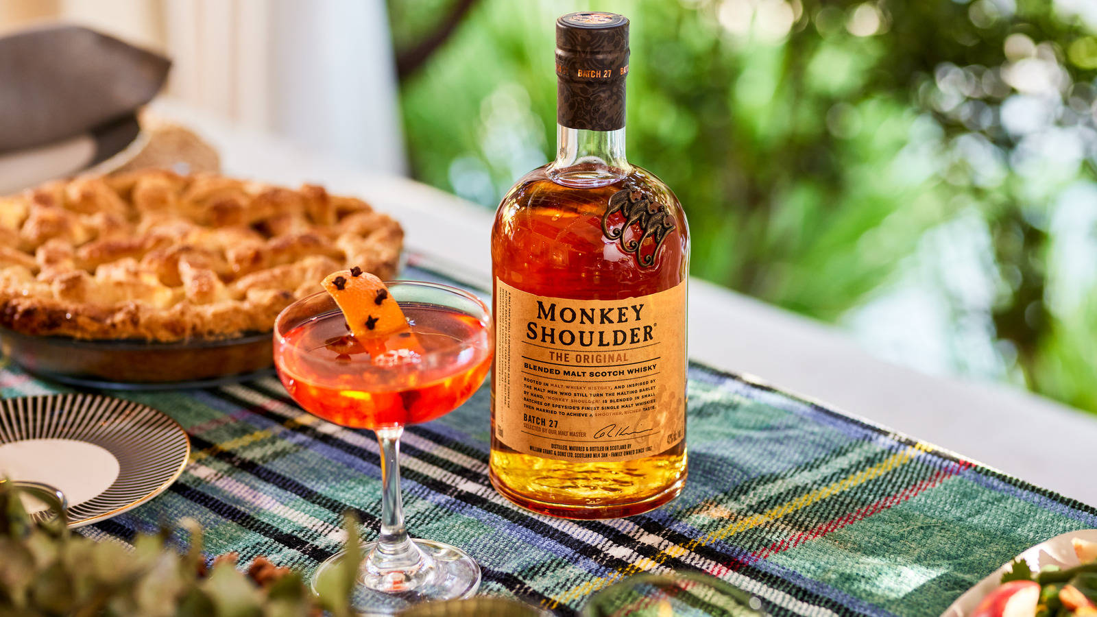 alcohol 桌布 壁紙 — 一瓶Monkey Shoulder威士忌和一杯以柳橙裝飾的雞尾酒，放在戶外格子桌布上的一個金黃色派旁邊。