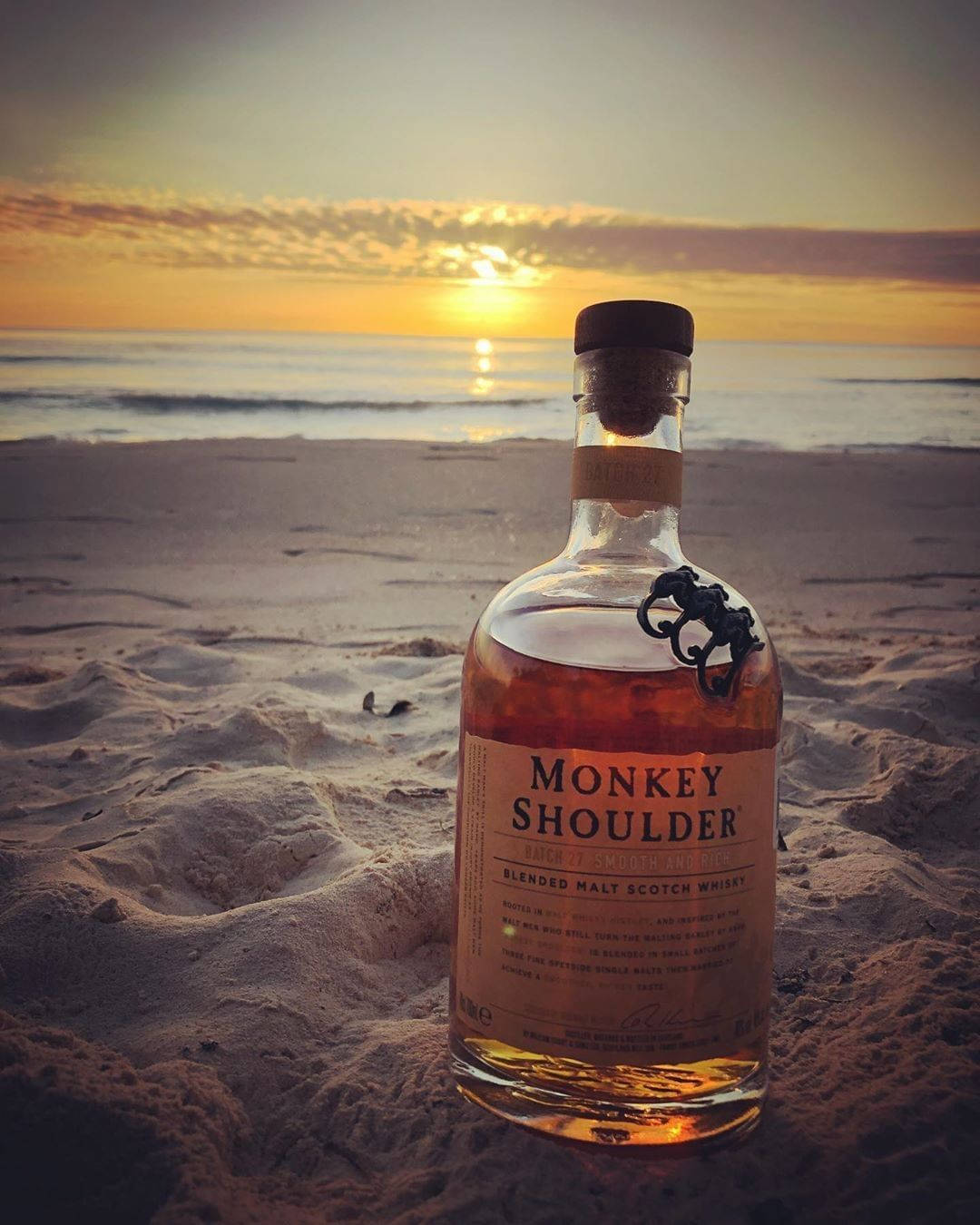 飲料 桌布 壁紙 — 一瓶Monkey Shoulder威士忌放在溫暖的沙上，夕陽在海面上落下。