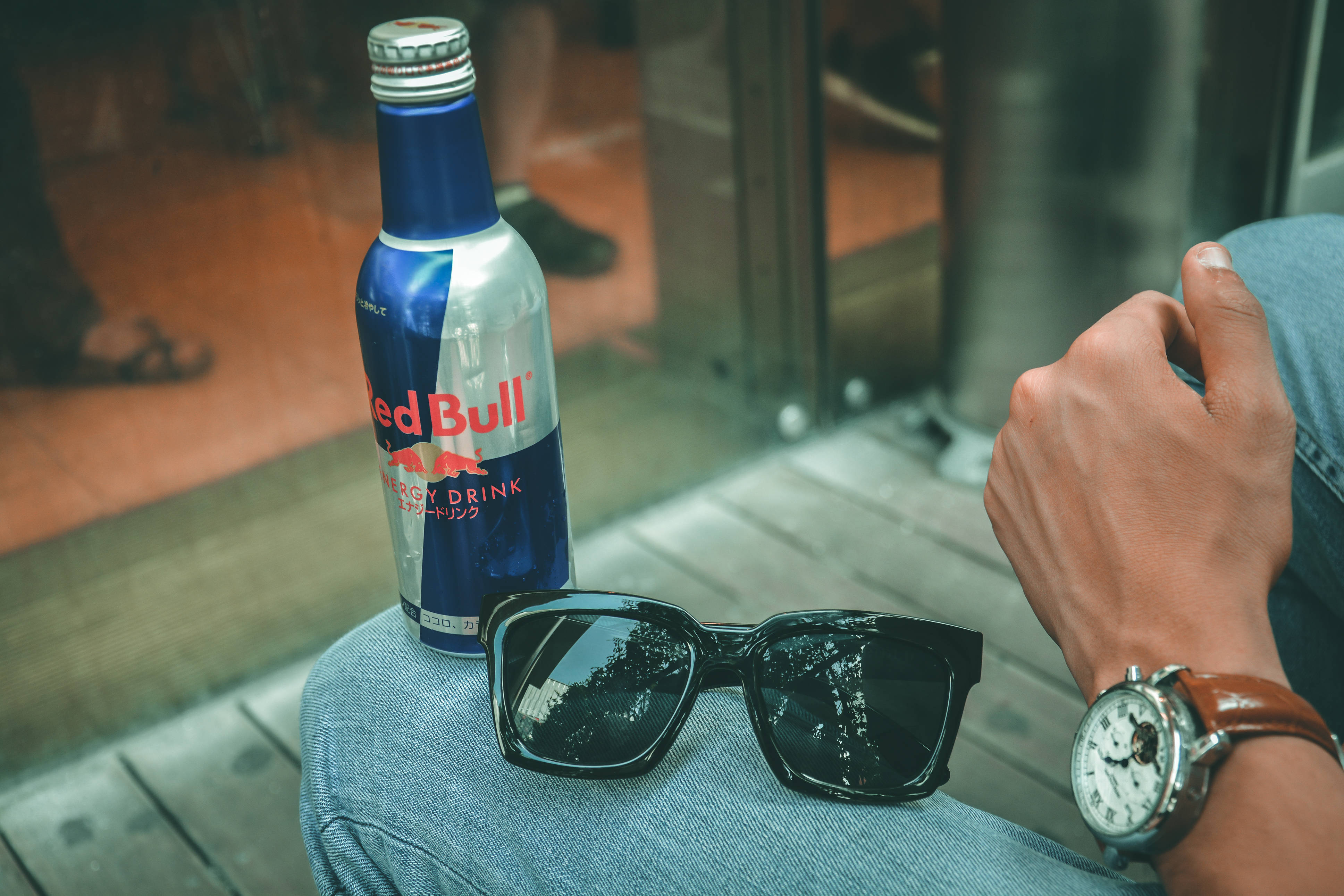 bull 桌布 壁紙 — 一瓶Red Bull能量飲料、太陽眼鏡和手錶放在靠近玻璃門的牛仔褲腿上。