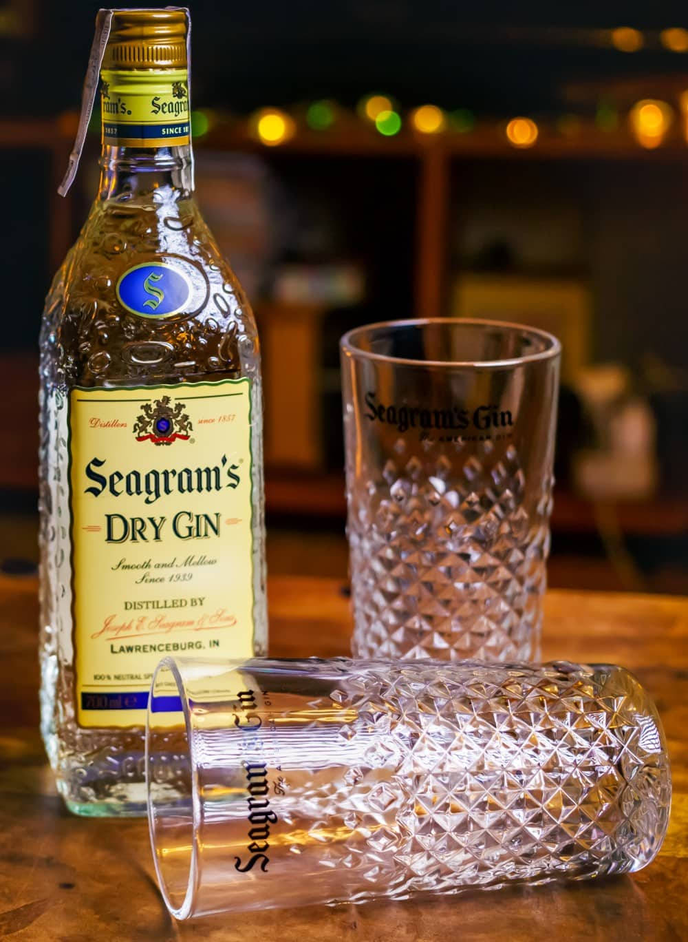 高清 桌布 壁紙 — 一瓶Seagram's Dry Gin和兩個帶圖案的玻璃杯放在木製吧檯上，背景有溫暖的散景燈光。