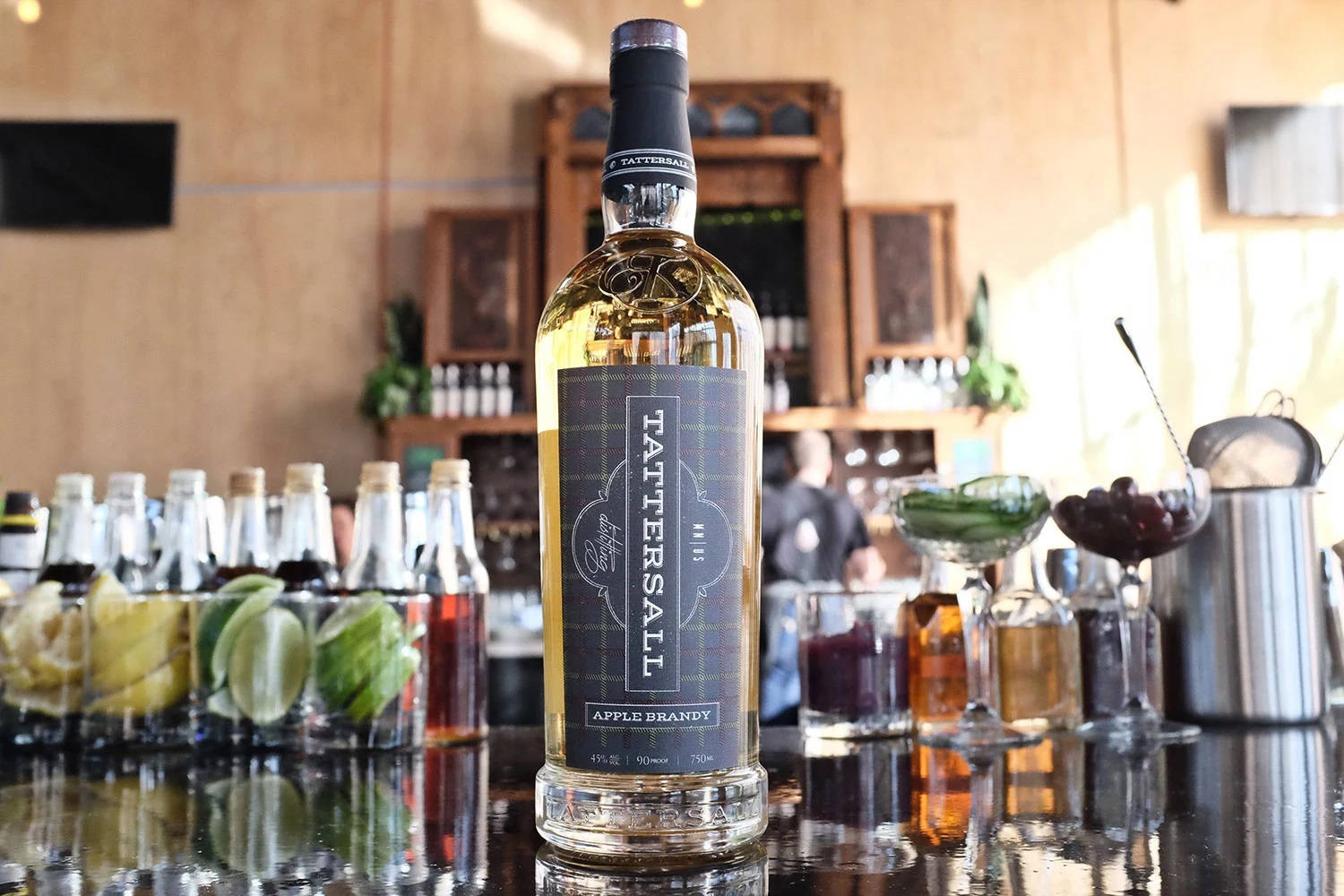 飲料 桌布 壁紙 — 一瓶Tattersall Apple Brandy在拋光吧檯上閃耀，周圍環繞著雞尾酒杯和柑橘裝飾。