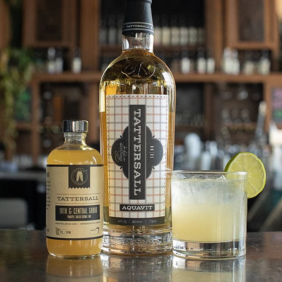 飲料 桌布 壁紙 — 一瓶Tattersall Aquavit和一瓶較小的10th & Central Shrub放在吧檯上，旁邊有一杯以萊姆裝飾的雞尾酒。