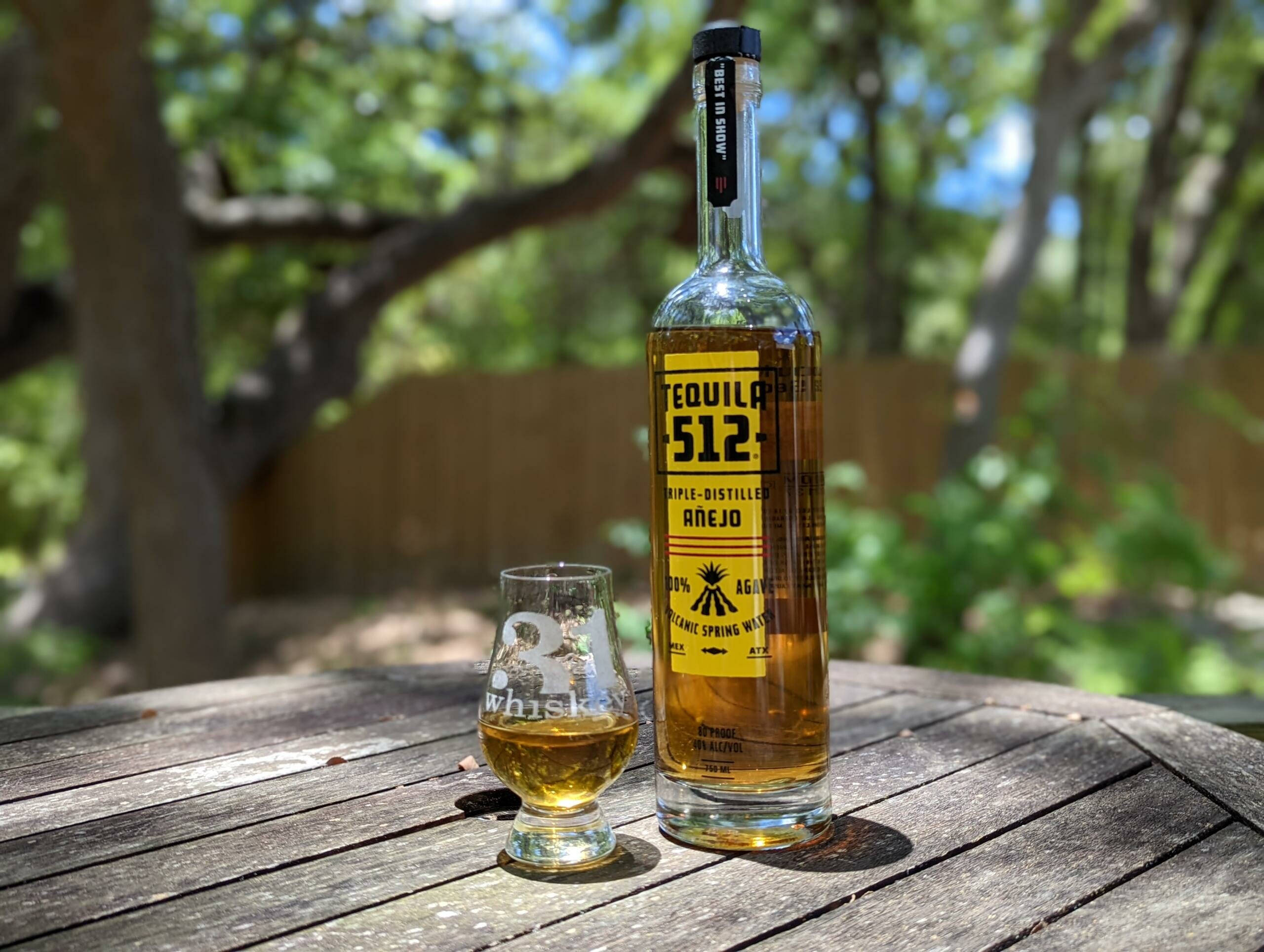 drinks 桌布 壁紙 — 一瓶Tequila 512 Añejo和一個裝滿琥珀色液體的品酒杯放在戶外一張風化的木桌上，背景有樹木，陽光普照。