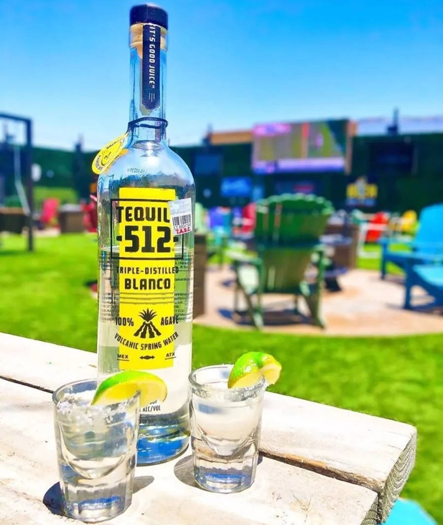 drinks 桌布 壁紙 — 一瓶Tequila -512 Blanco和兩杯以萊姆裝飾的烈酒，放在戶外酒吧的木桌上，背景是明亮的藍天。