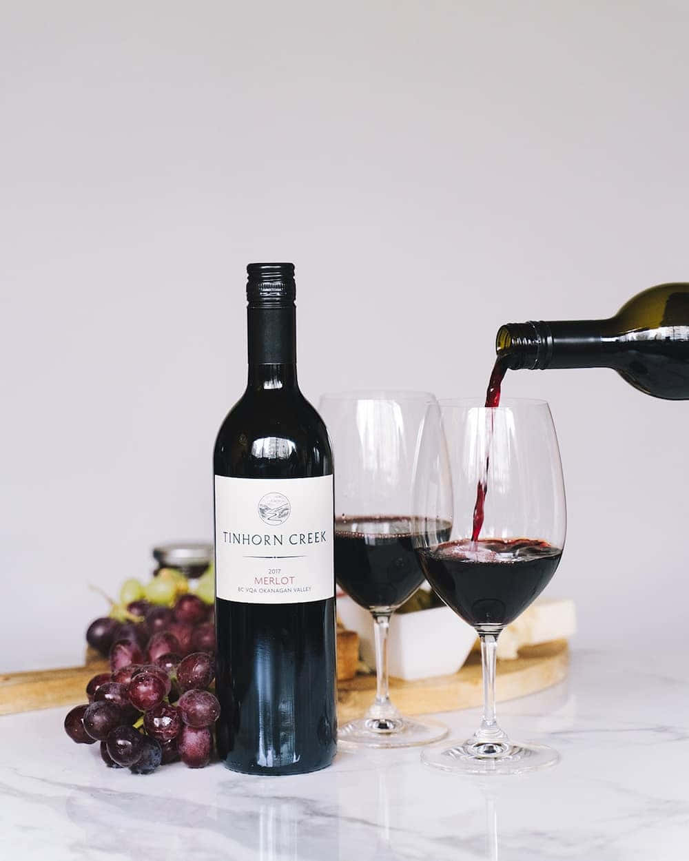 Black Wine 桌布 壁紙 — 一瓶Tinhorn Creek Merlot在大理石表面上將濃郁的紅酒倒入酒杯中，旁邊放著新鮮葡萄。