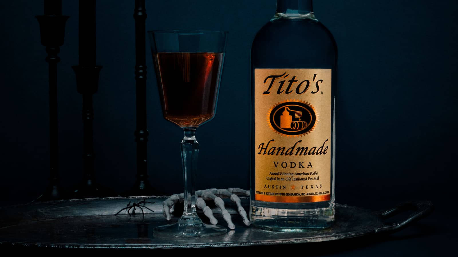 glass 桌布 壁紙 — 一瓶Tito's Handmade Vodka和一杯琥珀色液體放在托盤上，旁邊有一隻骷髏手，背景是深藍色。