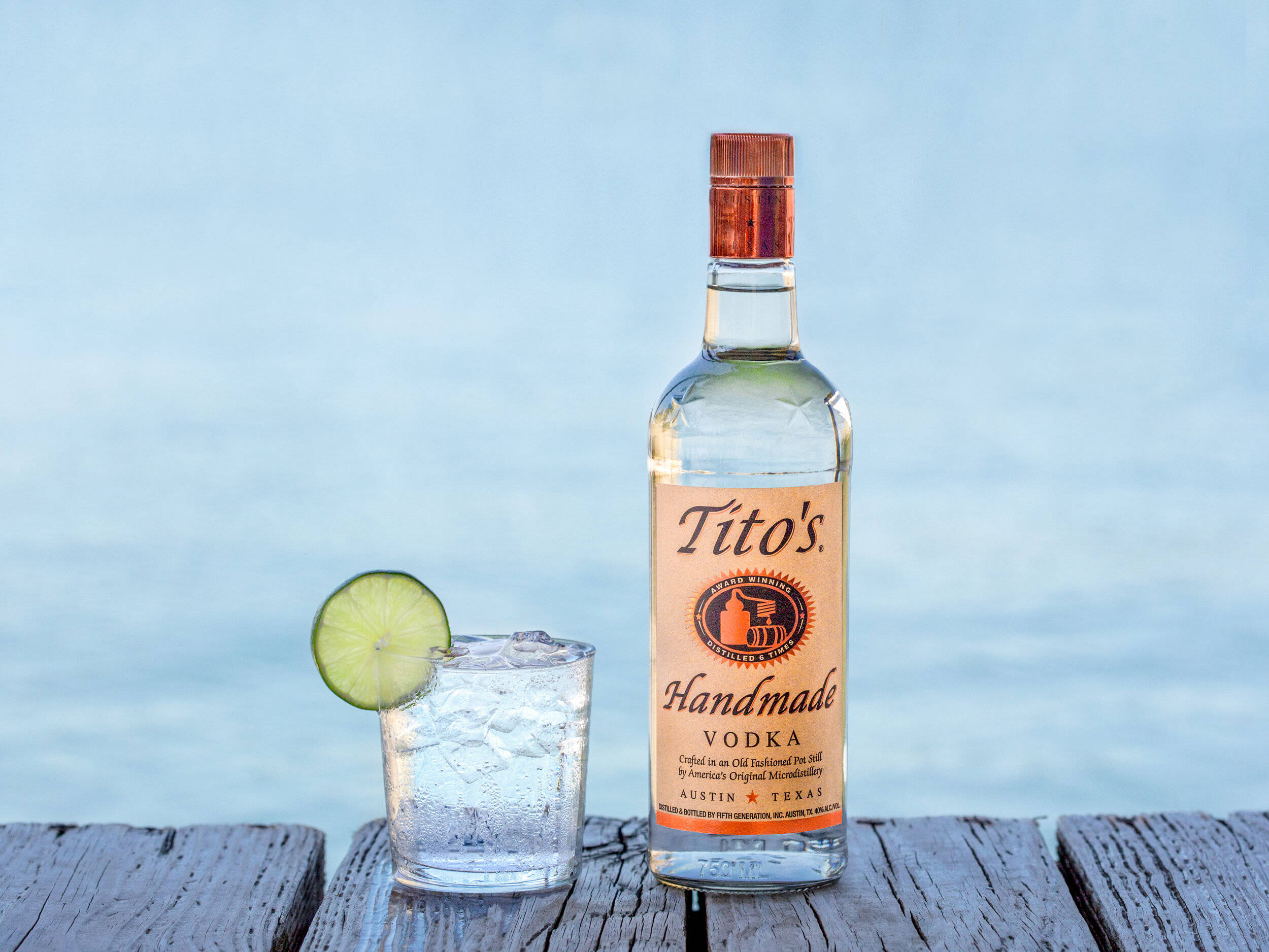glass 桌布 壁紙 — 一瓶Tito's Handmade Vodka和一杯以萊姆裝飾的雞尾酒，放在一條經風化的木碼頭上，背景是平靜的藍色水面。