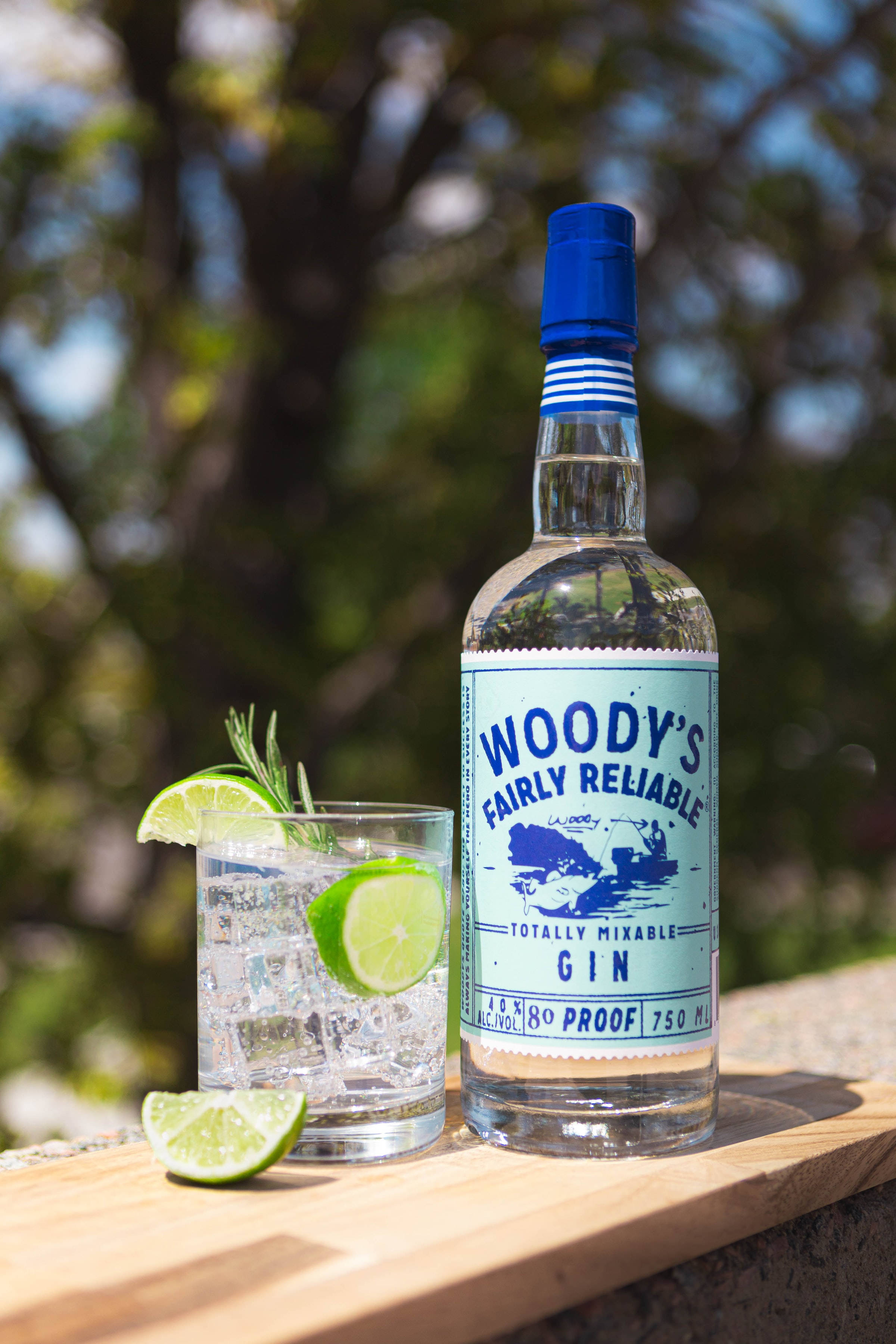 drinks 桌布 壁紙 — 一瓶Woody's Fairly Reliable Gin放在戶外木質表面上，旁邊是一杯以青檸和迷迭香裝飾的琴酒雞尾酒。