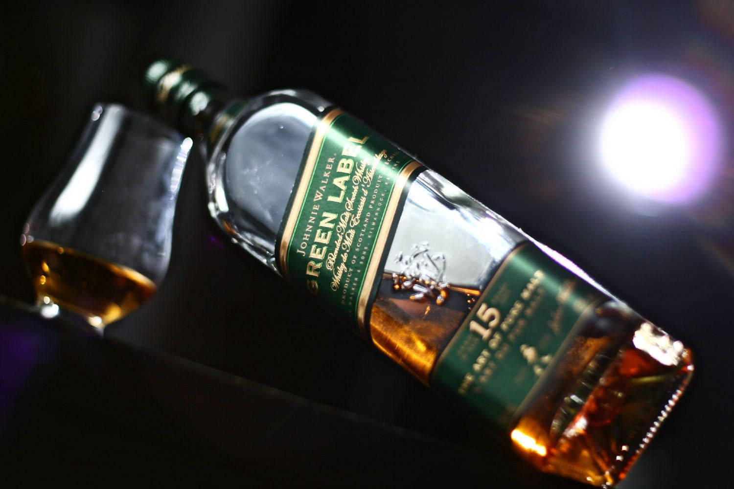 alcohol 桌布 壁紙 — 一瓶傾斜的Johnnie Walker Green Label威士忌，旁邊放著一個玻璃杯，在黑暗背景下被明亮的紫色鏡頭光暈照亮。