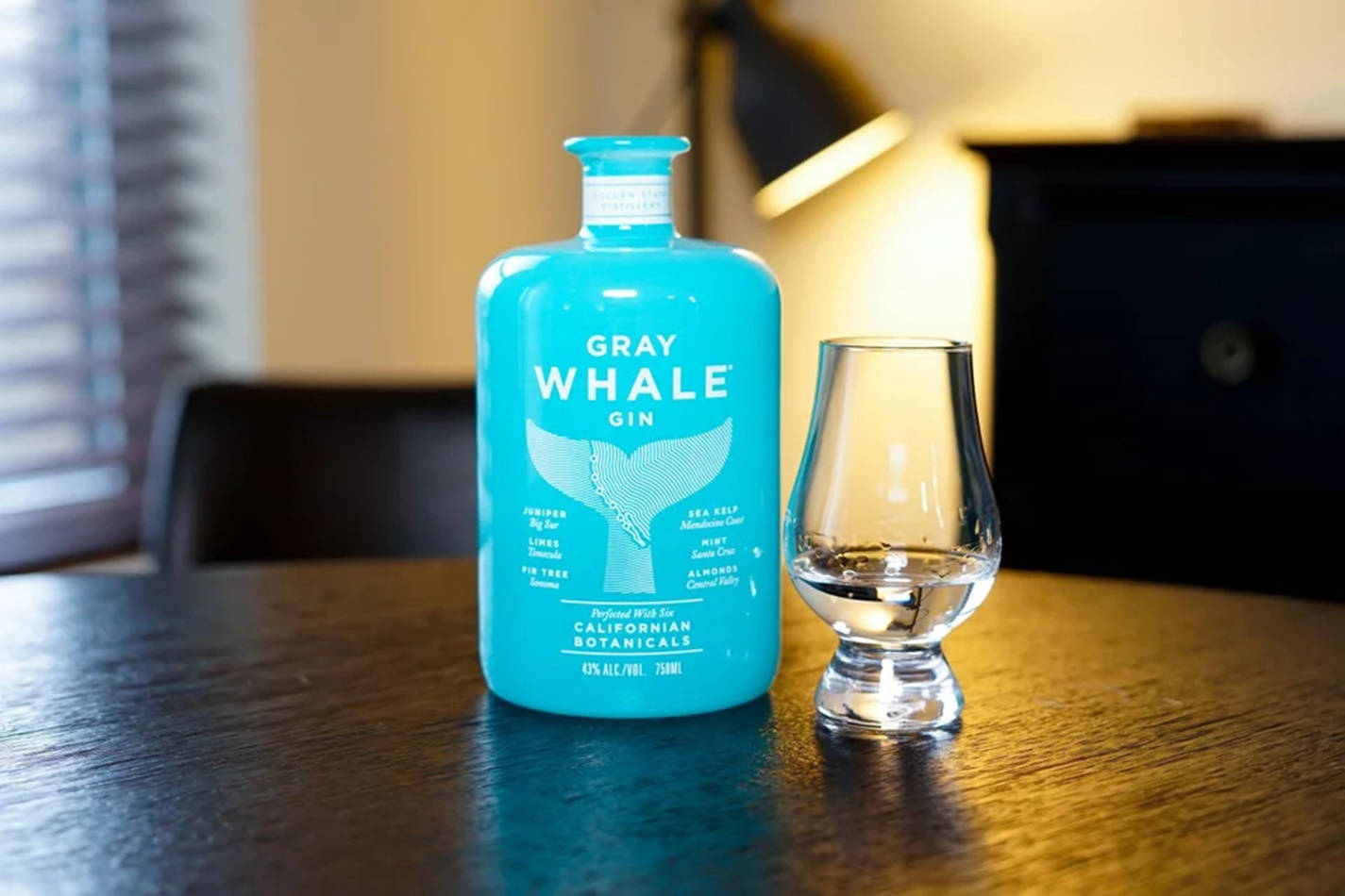 Whale 桌布 壁紙 — 一瓶充滿活力的藍綠色Gray Whale Gin酒瓶放在一張溫暖的木桌上，旁邊有一個空的品酒杯，房間光線柔和。