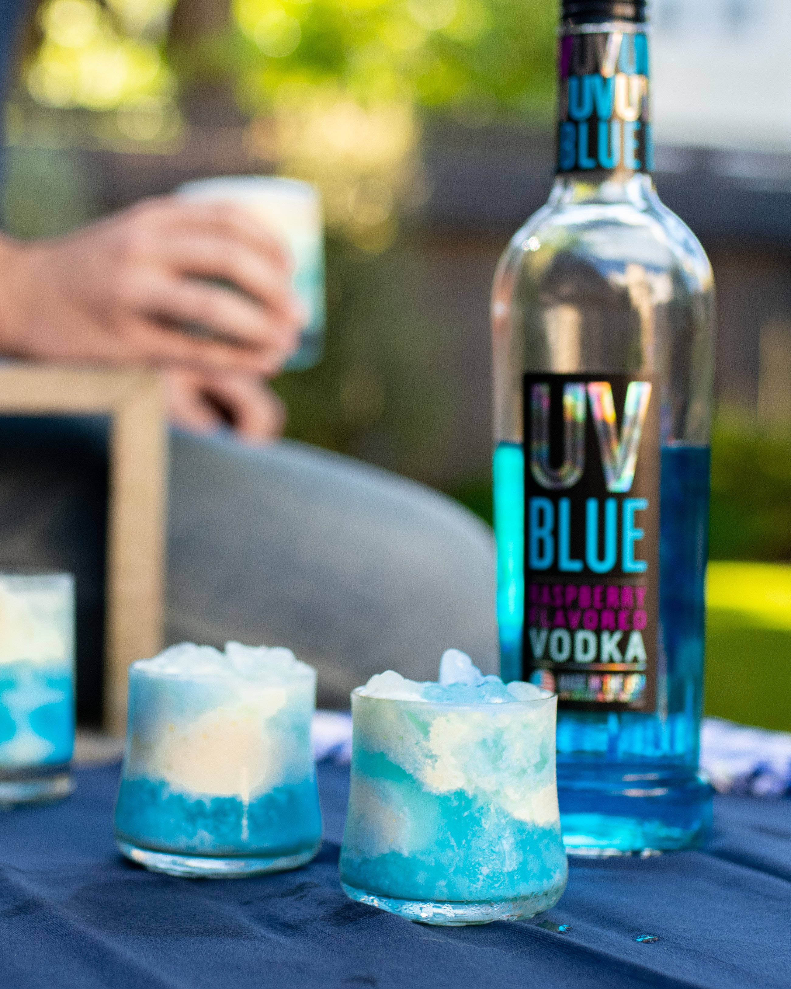 drinks 桌布 壁紙 — 一瓶充滿活力的藍色覆盆子伏特加酒和分層雞尾酒放在戶外的深色桌布上，背景中有人在放鬆。