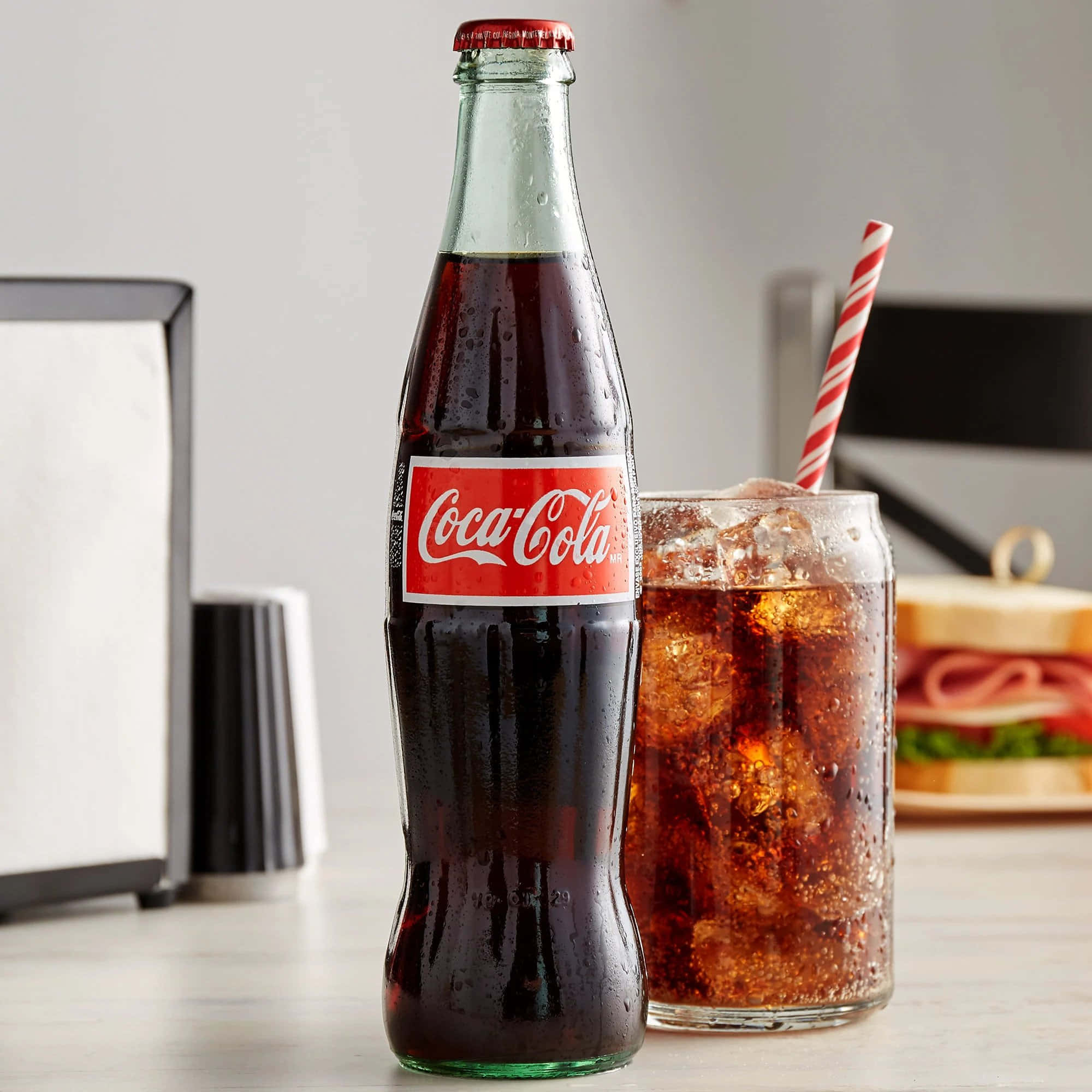 coca cola 桌布 壁紙 — 一瓶冰凍玻璃瓶裝可口可樂和一杯加冰及條紋吸管的可口可樂，放在乾淨的檯面上，旁邊有一個三明治。