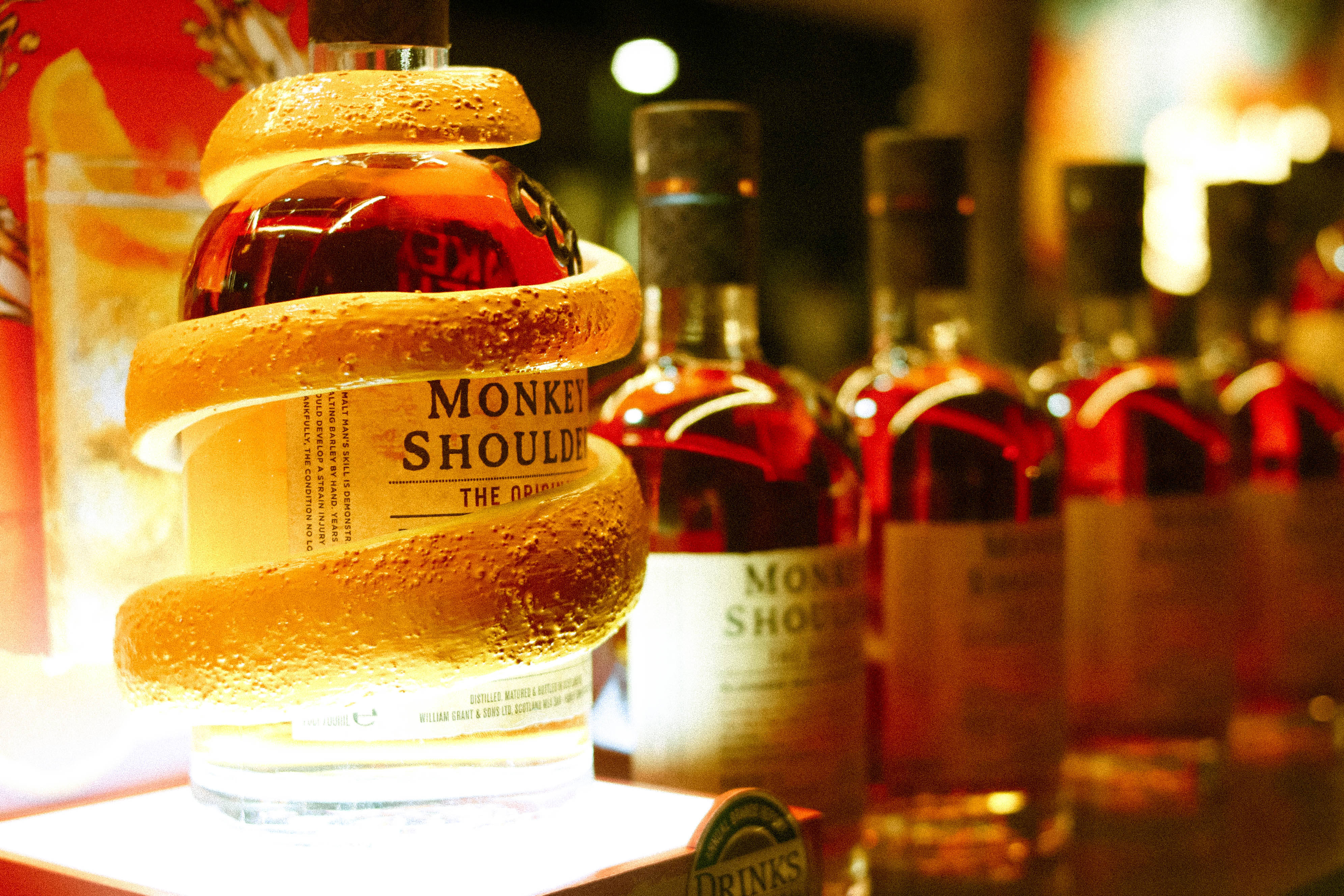 alcohol 桌布 壁紙 — 一瓶冰凍的Monkey Shoulder威士忌包裹在發光的柑橘皮中，在模糊的酒吧背景下溫暖地發光。