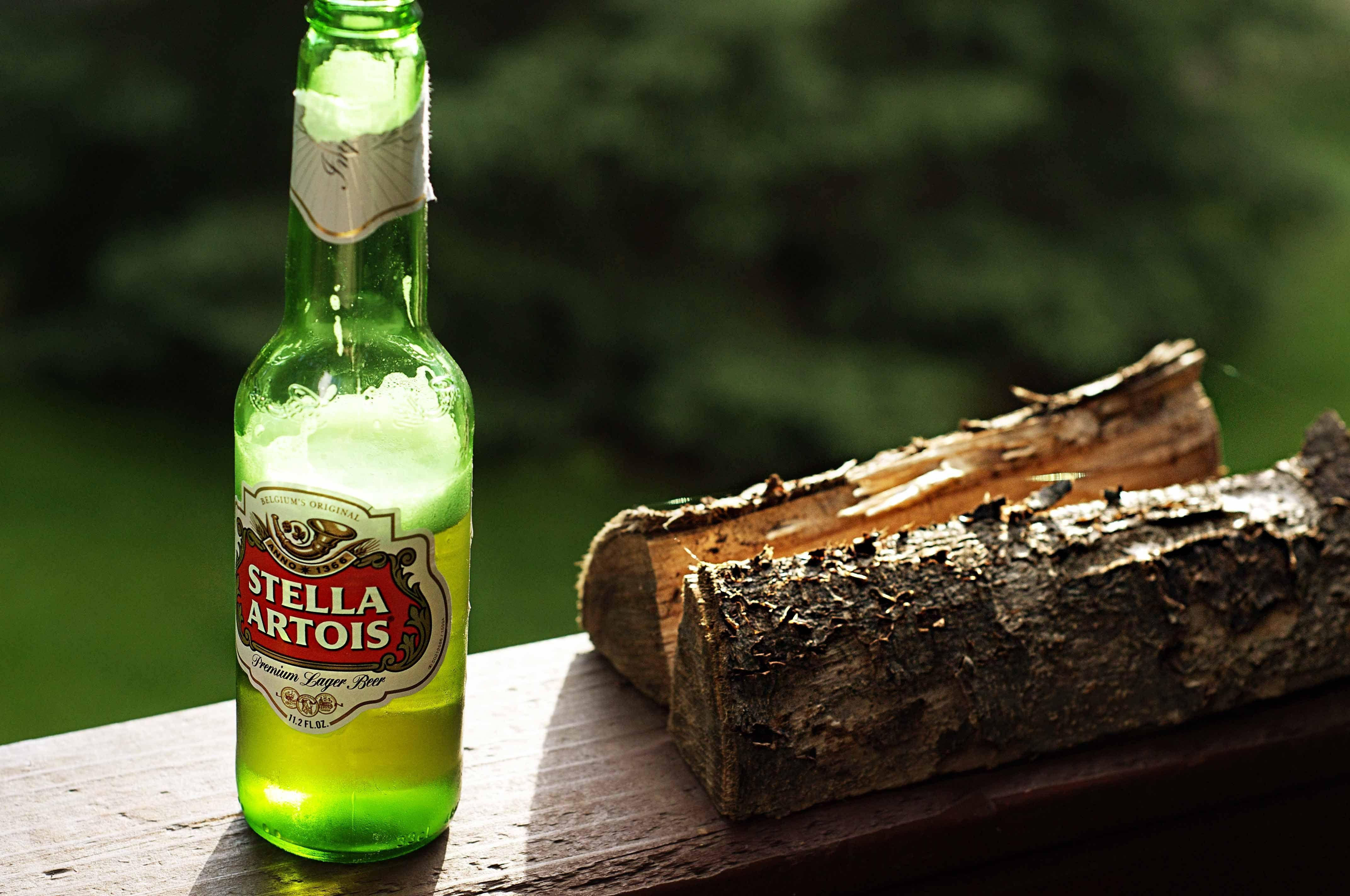 Beer 桌布 壁紙 — 一瓶冰涼的 Stella Artois 啤酒瓶，帶有冷凝水珠，放在木質甲板上，旁邊是粗切的木頭，背景是模糊的綠色森林。